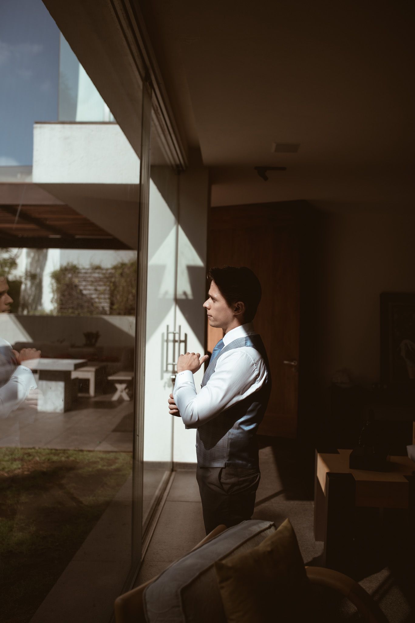 Person in formal attire gazes thoughtfully through large window in moody lighting.