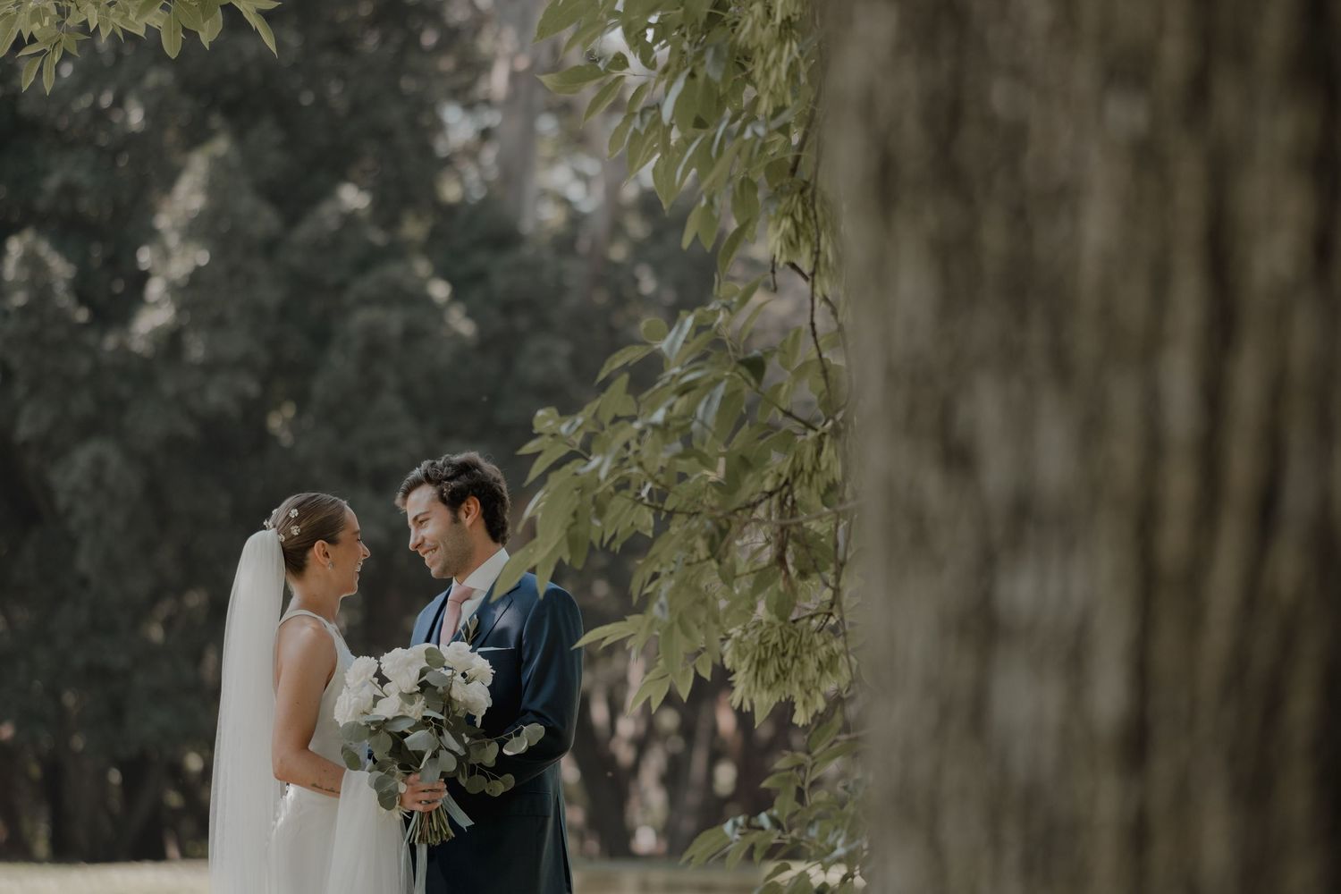 Newlyweds share an intimate moment under tree branches in a romantic outdoor setting.