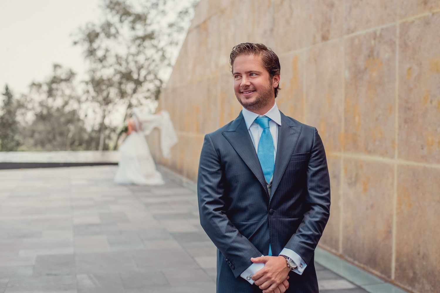 Elegant groom in a navy suit and turquoise tie stands against a stone wall.