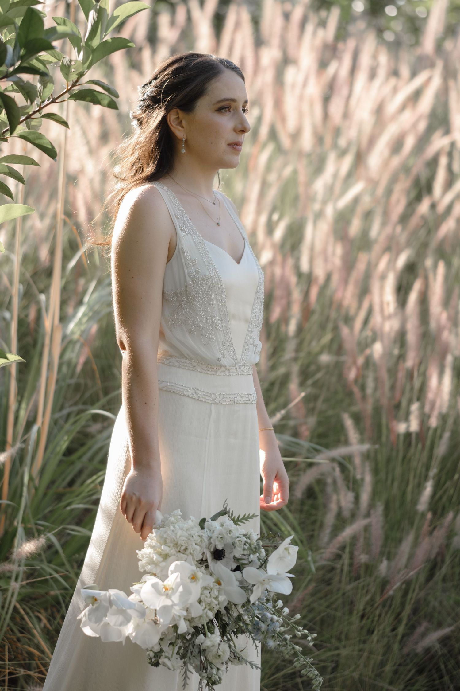 Bride in white dress stands in field of tall ornamental grasses holding white floral bouquet.