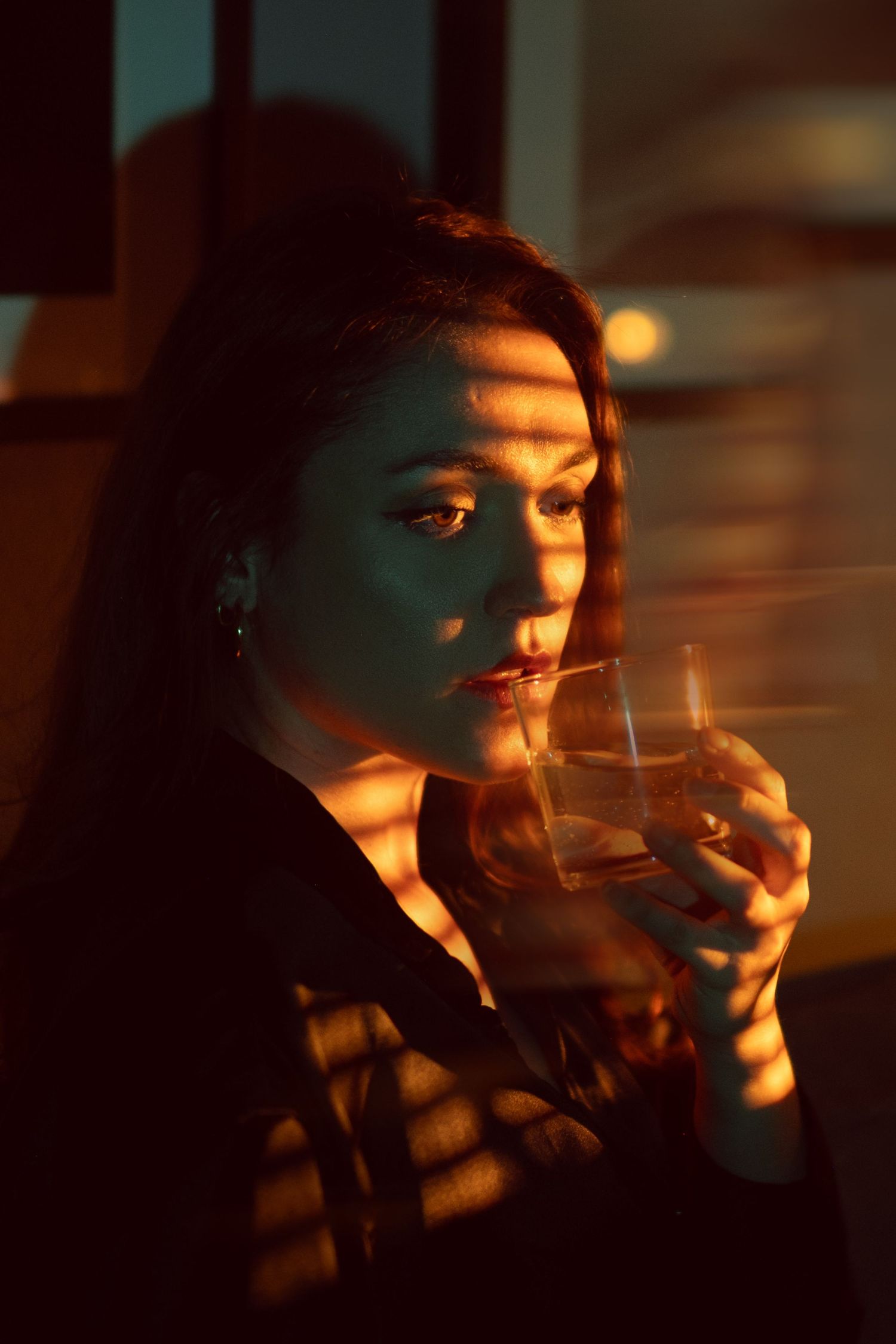 A dramatic portrait in warm orange lighting shows someone sipping from a glass as shadows create patterns across their face.