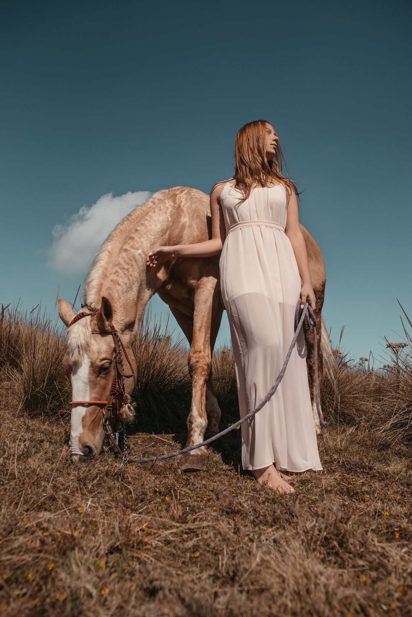 Woman in flowing white dress stands beside grazing palomino horse on grassy hillside.