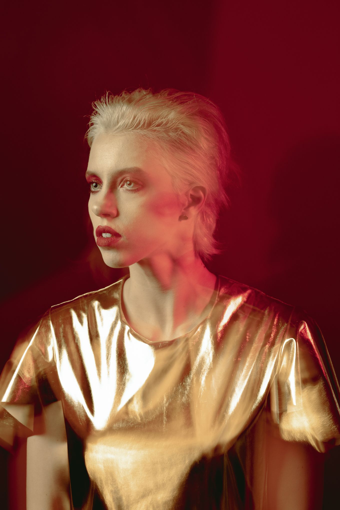 Model in metallic gold top poses against dark red backdrop with dramatic lighting and bold makeup.