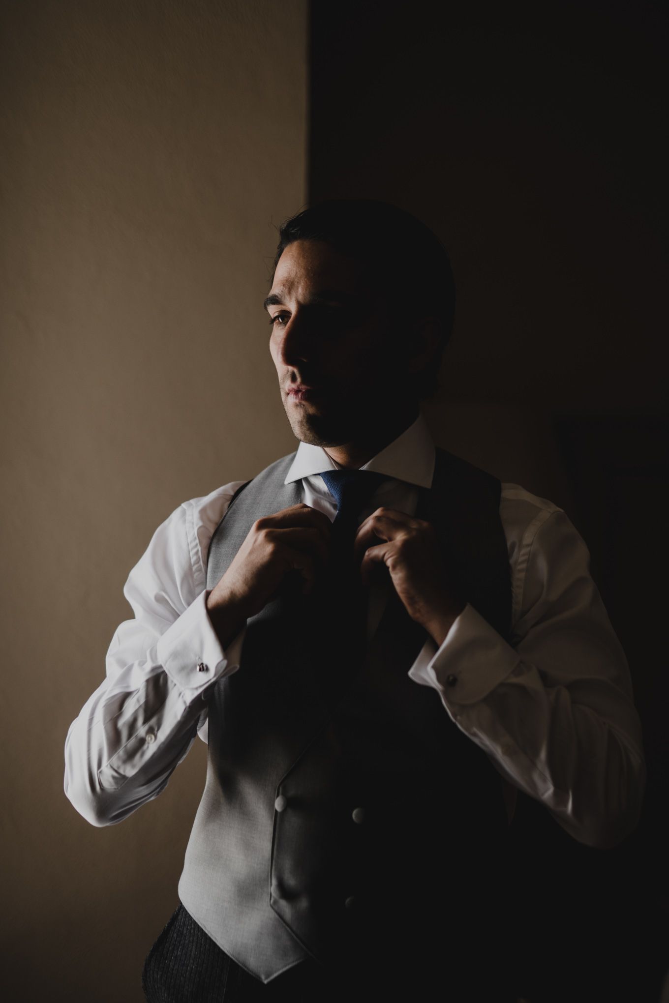 A person adjusts their tie while wearing a white dress shirt and gray vest in dramatic low-key lighting.