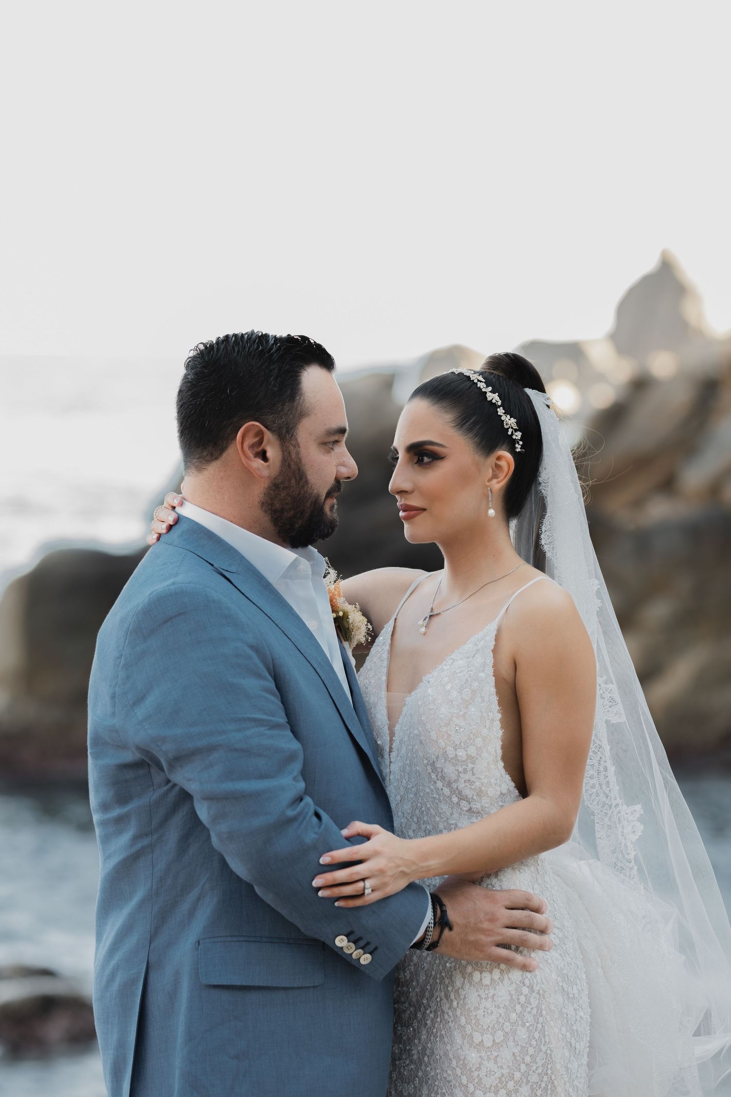 Newlywed couple in romantic pose by rocky ocean coast, bride in lace gown and groom in light blue suit.