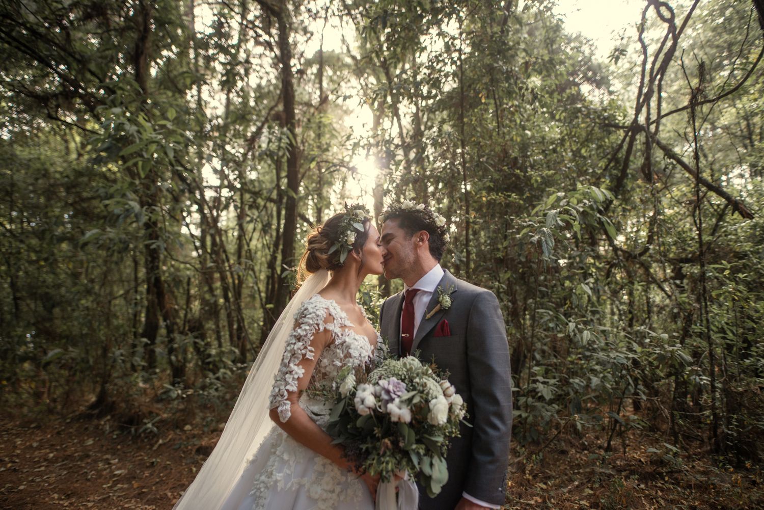A romantic wedding couple shares an intimate moment in a wooded forest setting with natural lighting and a white bouquet.