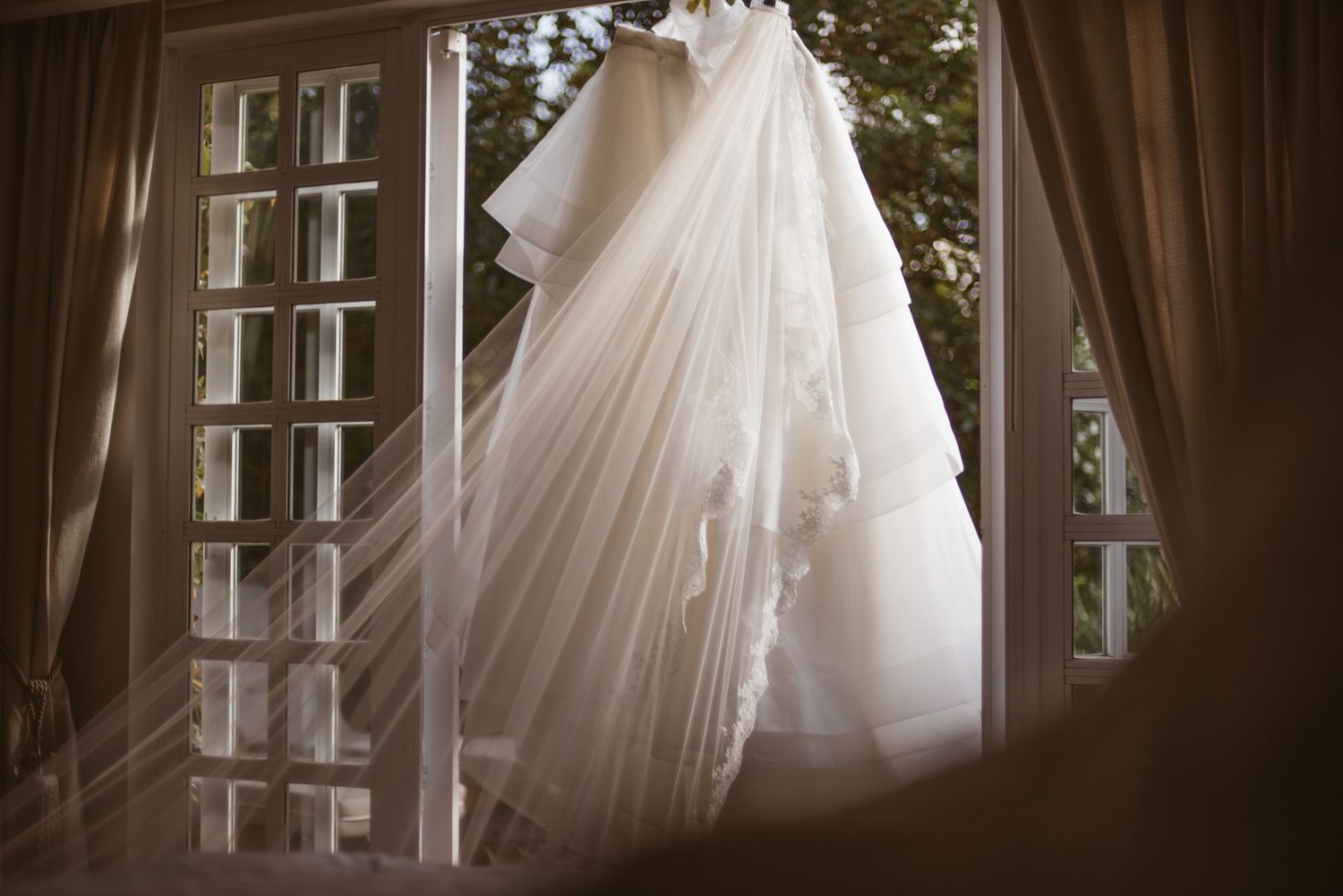 An elegant white wedding dress and veil hanging by a window with sheer curtains in soft natural lighting.