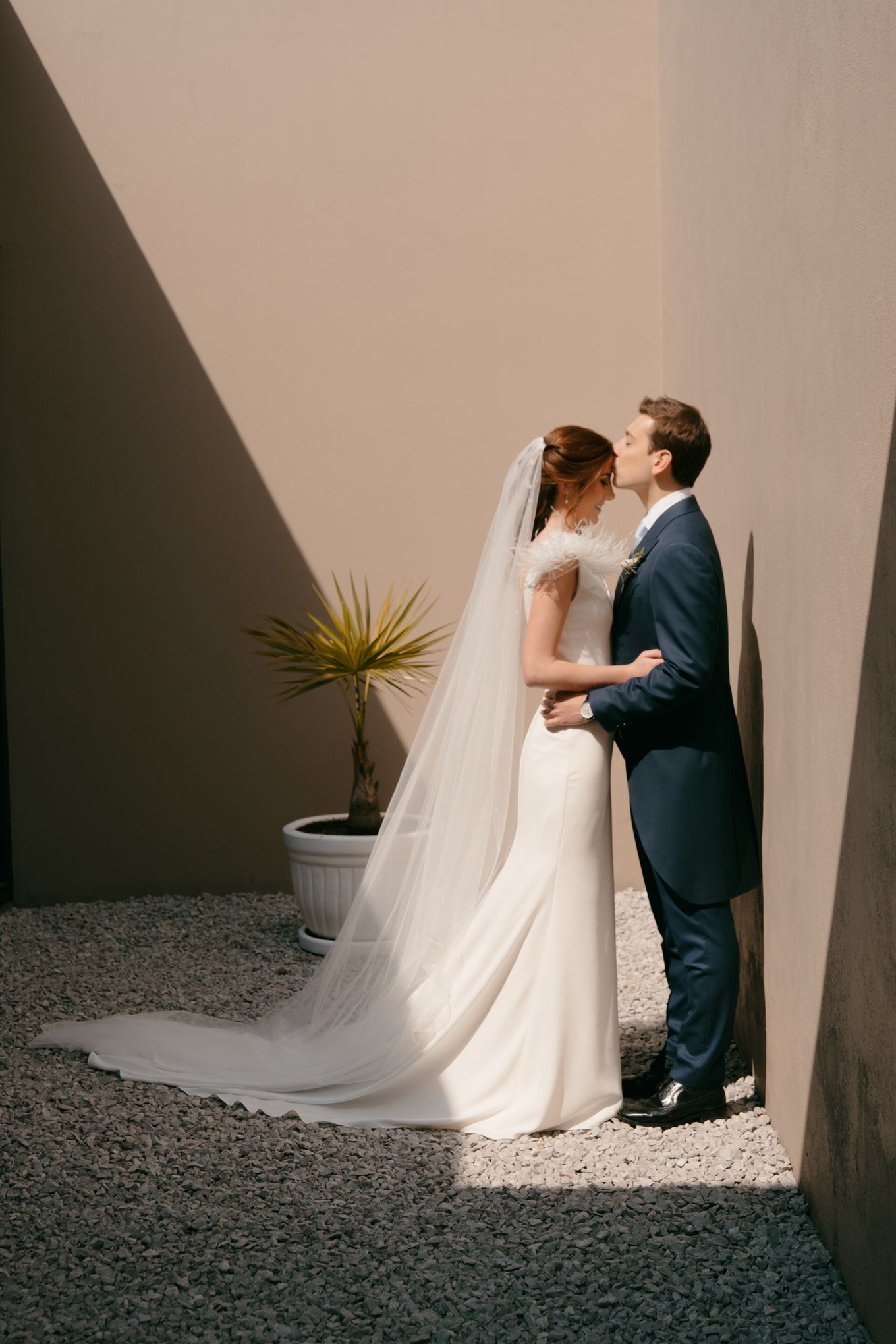 Wedding couple embraces in dramatic lighting between minimalist white walls.