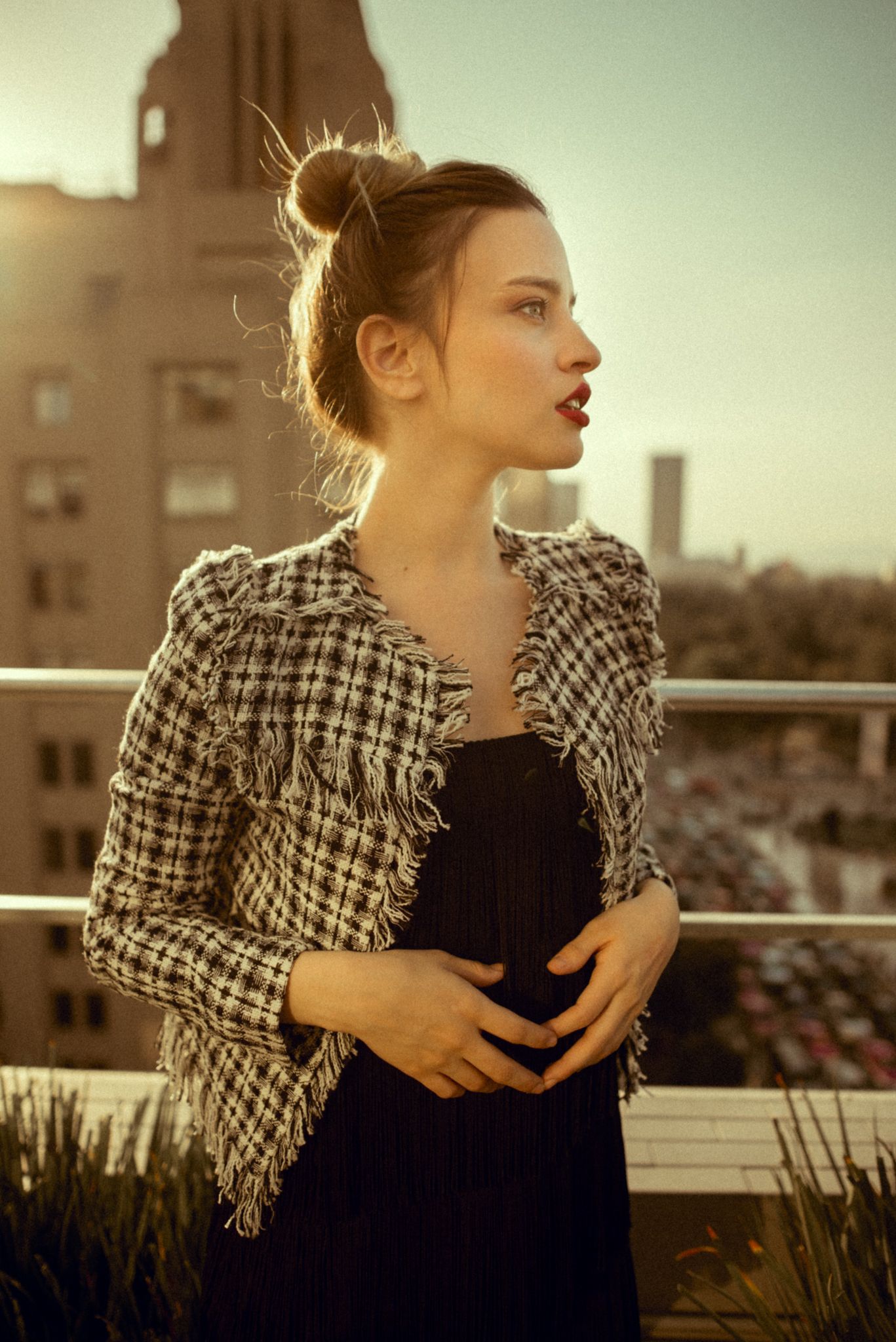 Vintage-inspired fashion shoot featuring plaid jacket and messy bun hairstyle against city skyline.