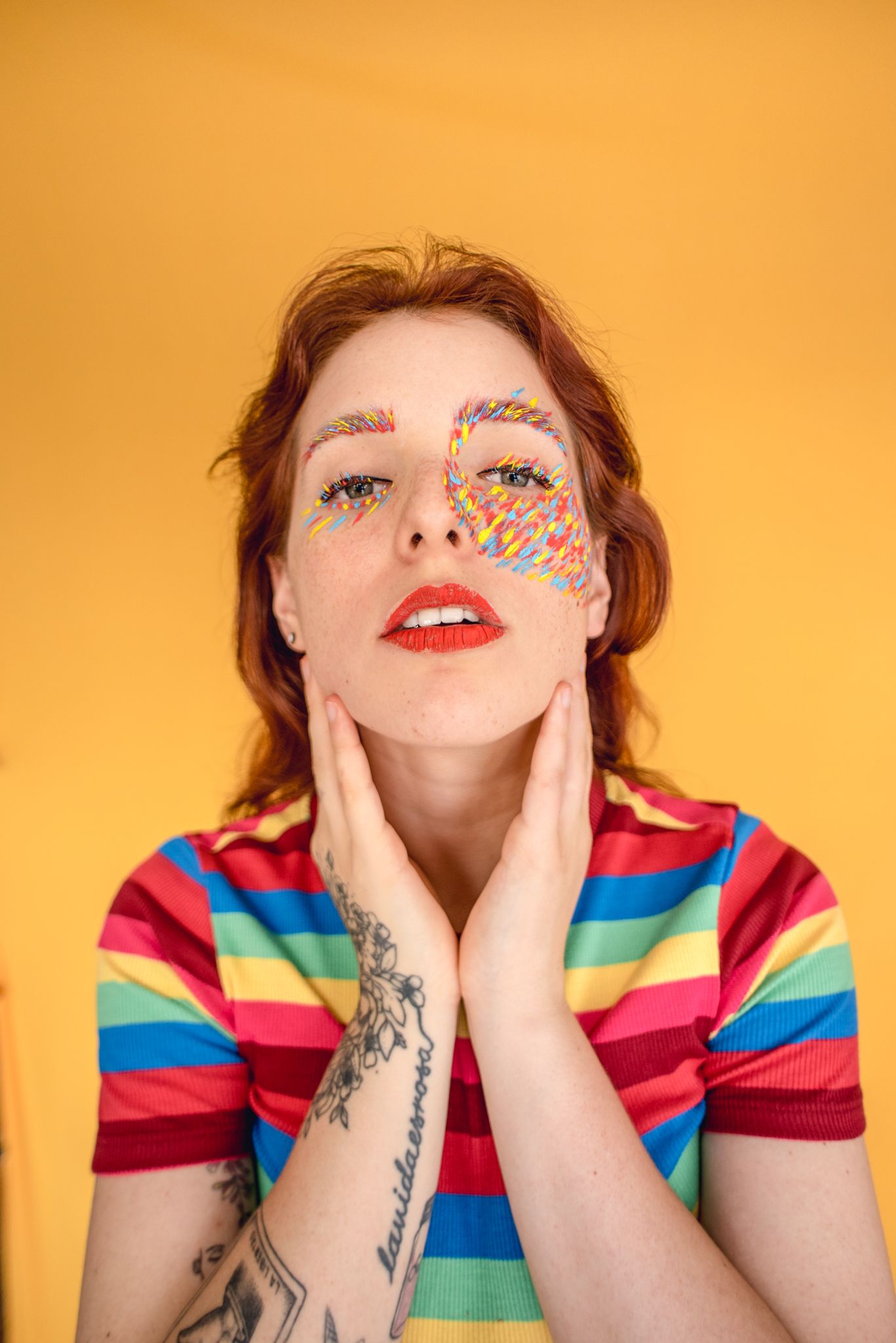 Colorful portrait with rainbow shirt and decorative face paint on orange backdrop.