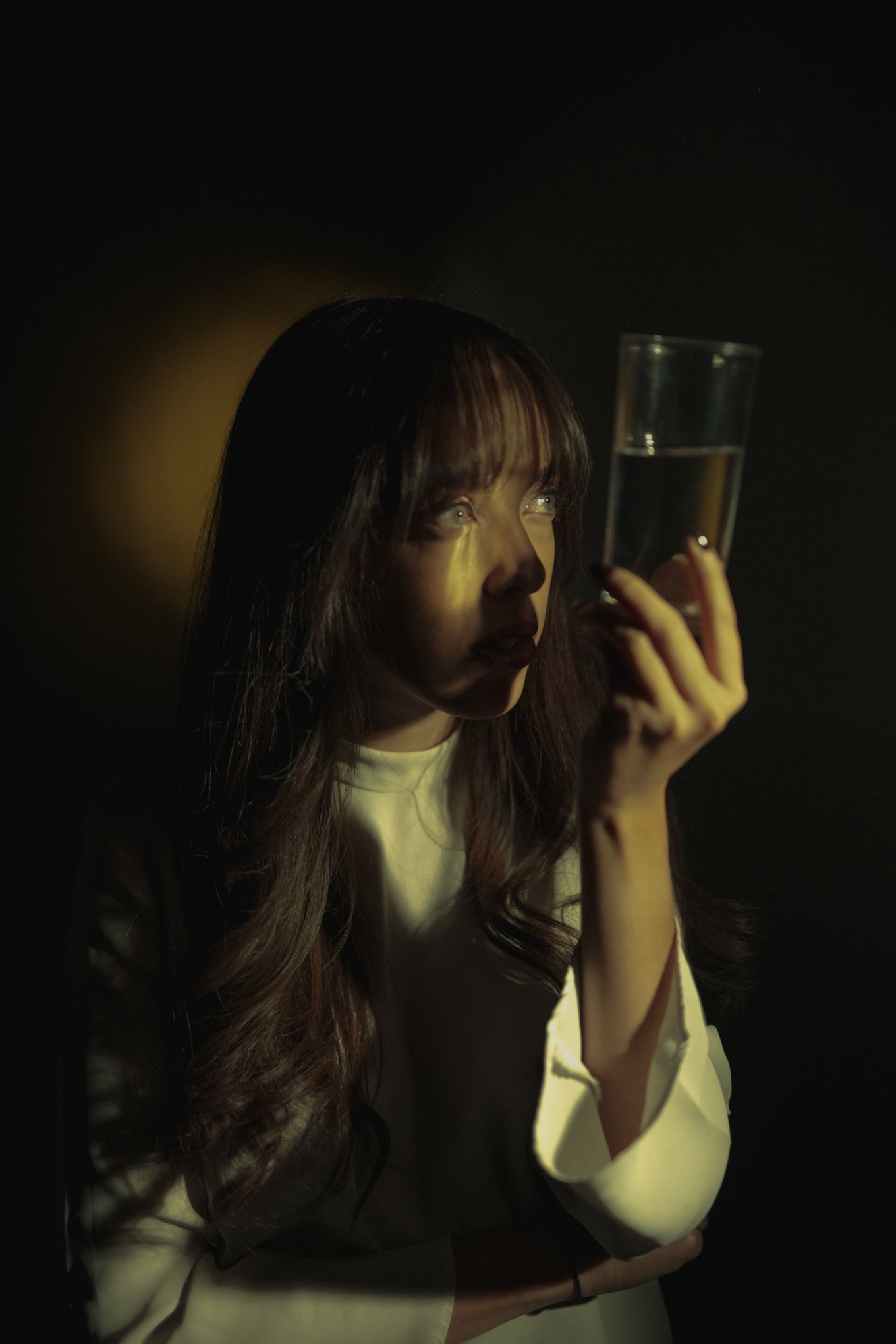Moody low-key portrait examining glass in dark setting.