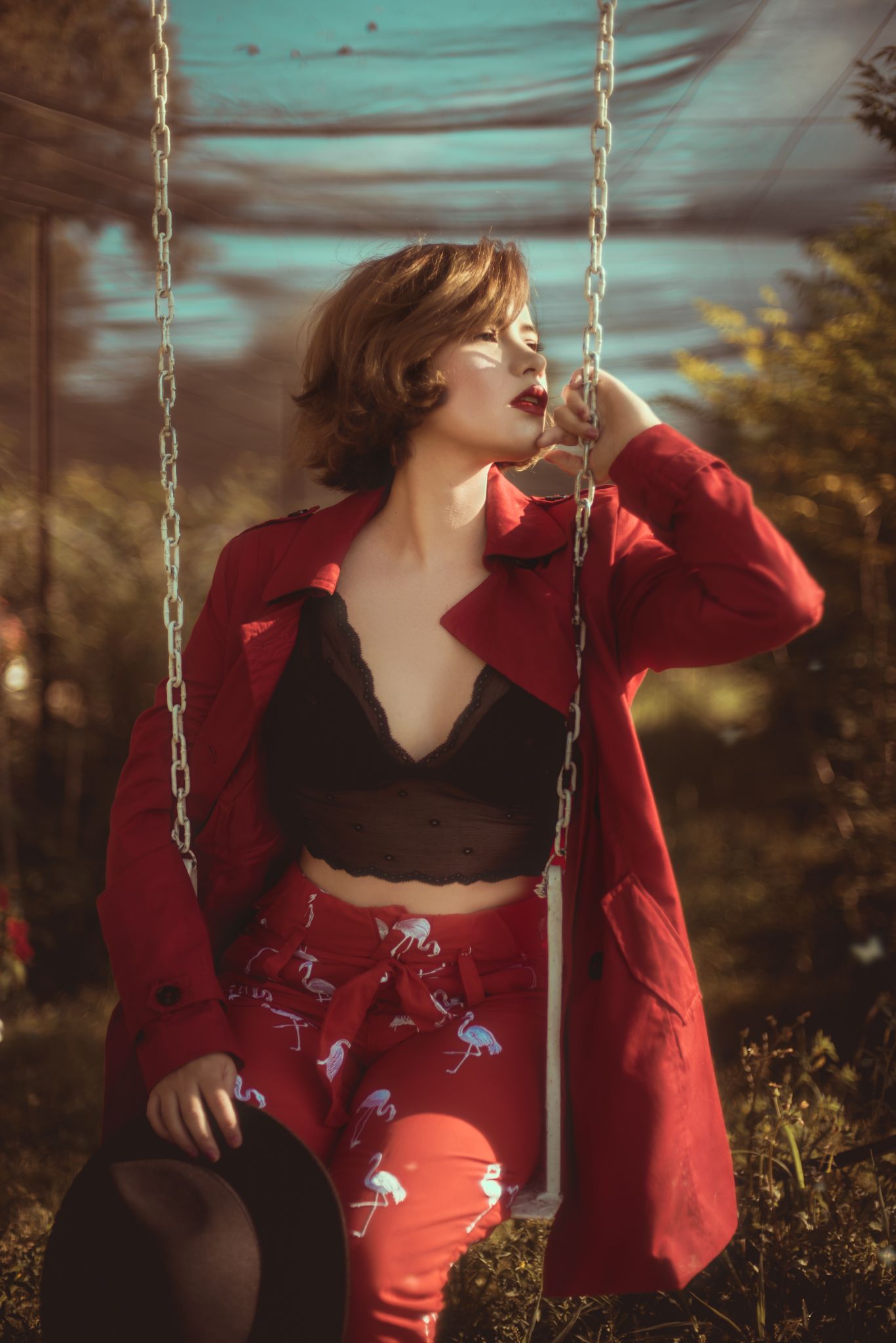 Stylish woman in red coat and black crop top sitting on swing in dreamy outdoor setting.