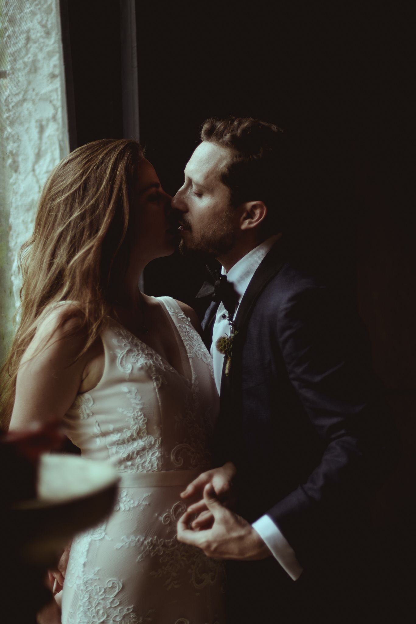 A romantic wedding moment with a couple sharing an intimate kiss in moody, dramatic lighting near a window.