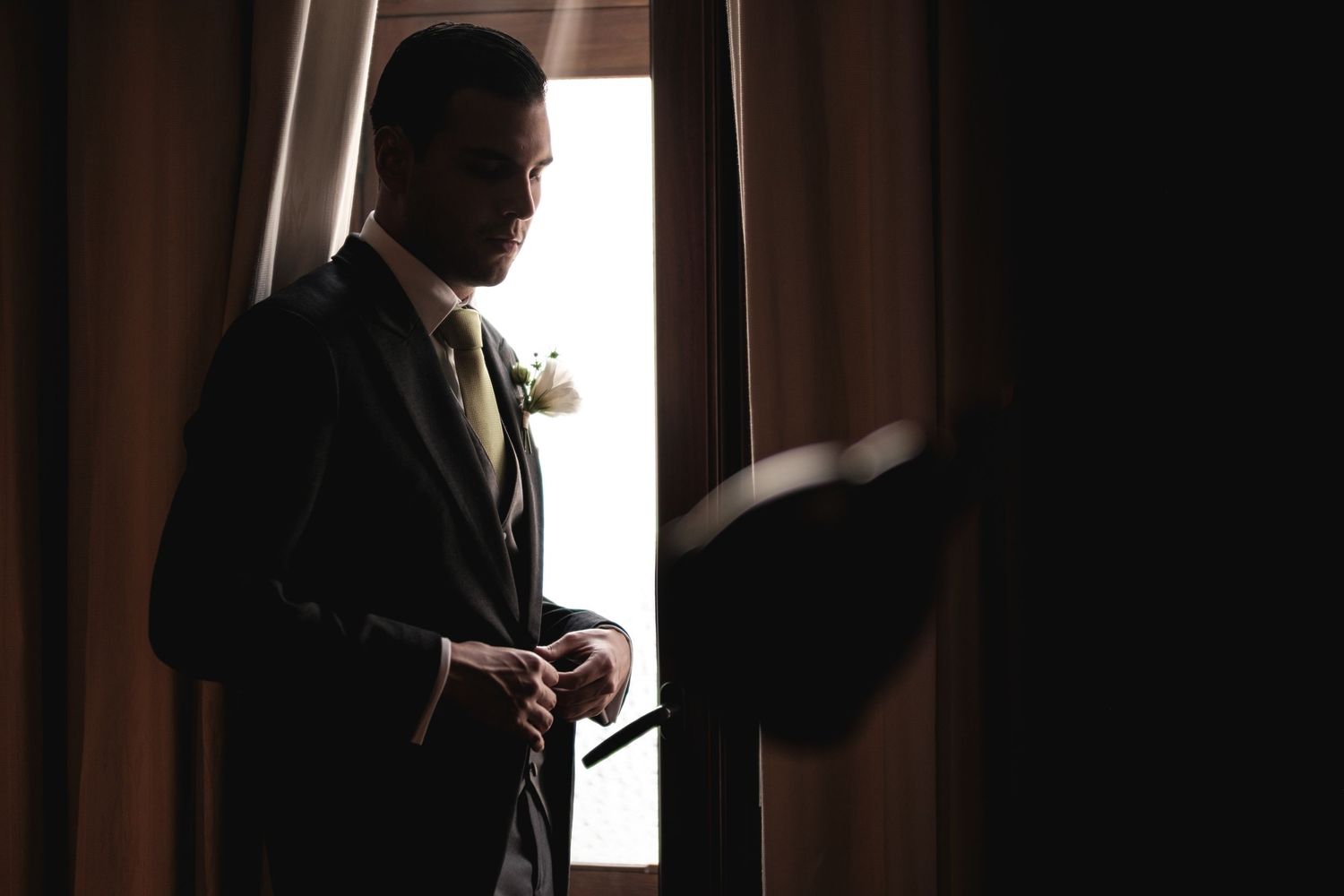 A silhouetted figure in a suit stands by a window with curtains in dramatic lighting.