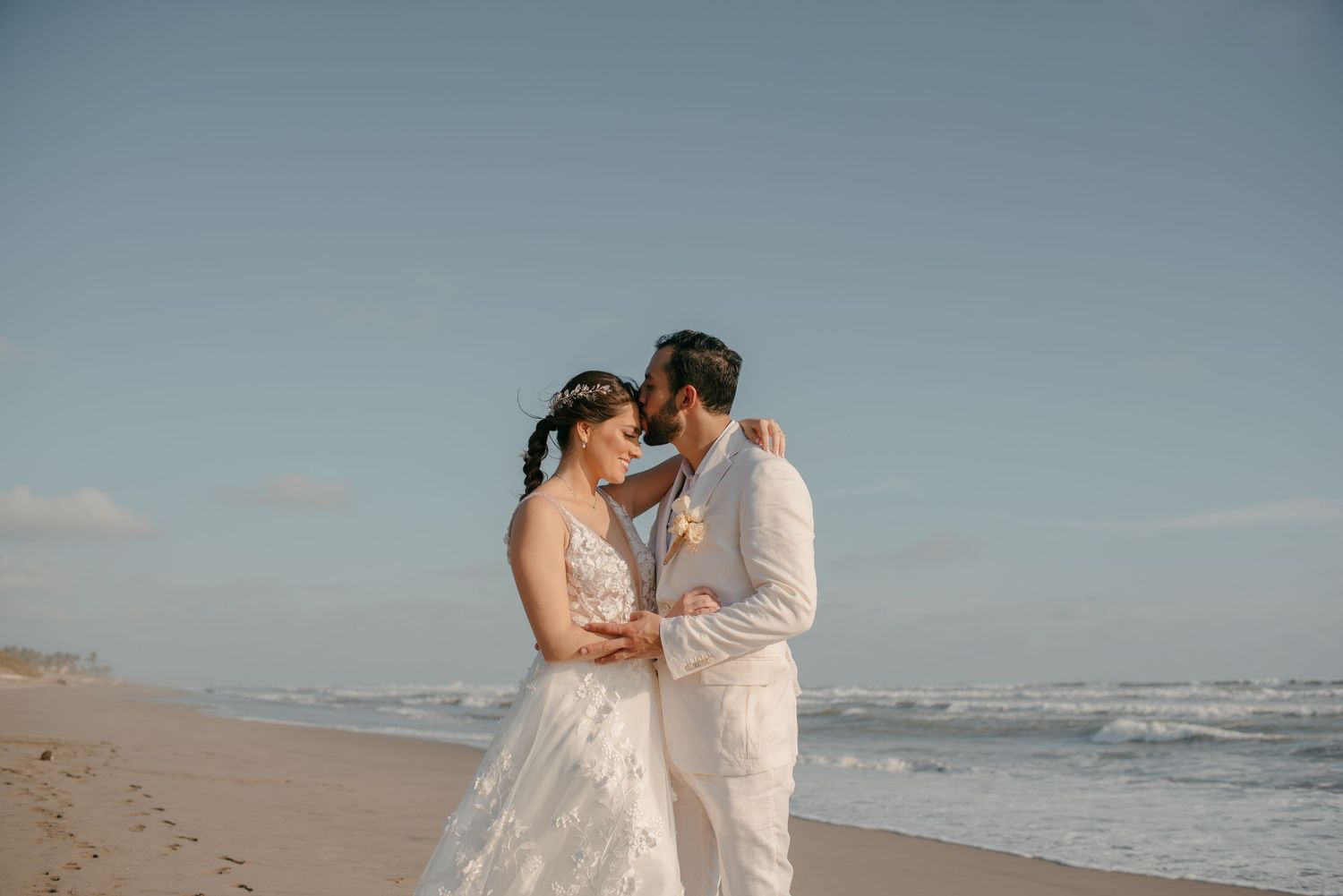 A couple in wedding attire embraces on a sandy beach with ocean waves in the background.