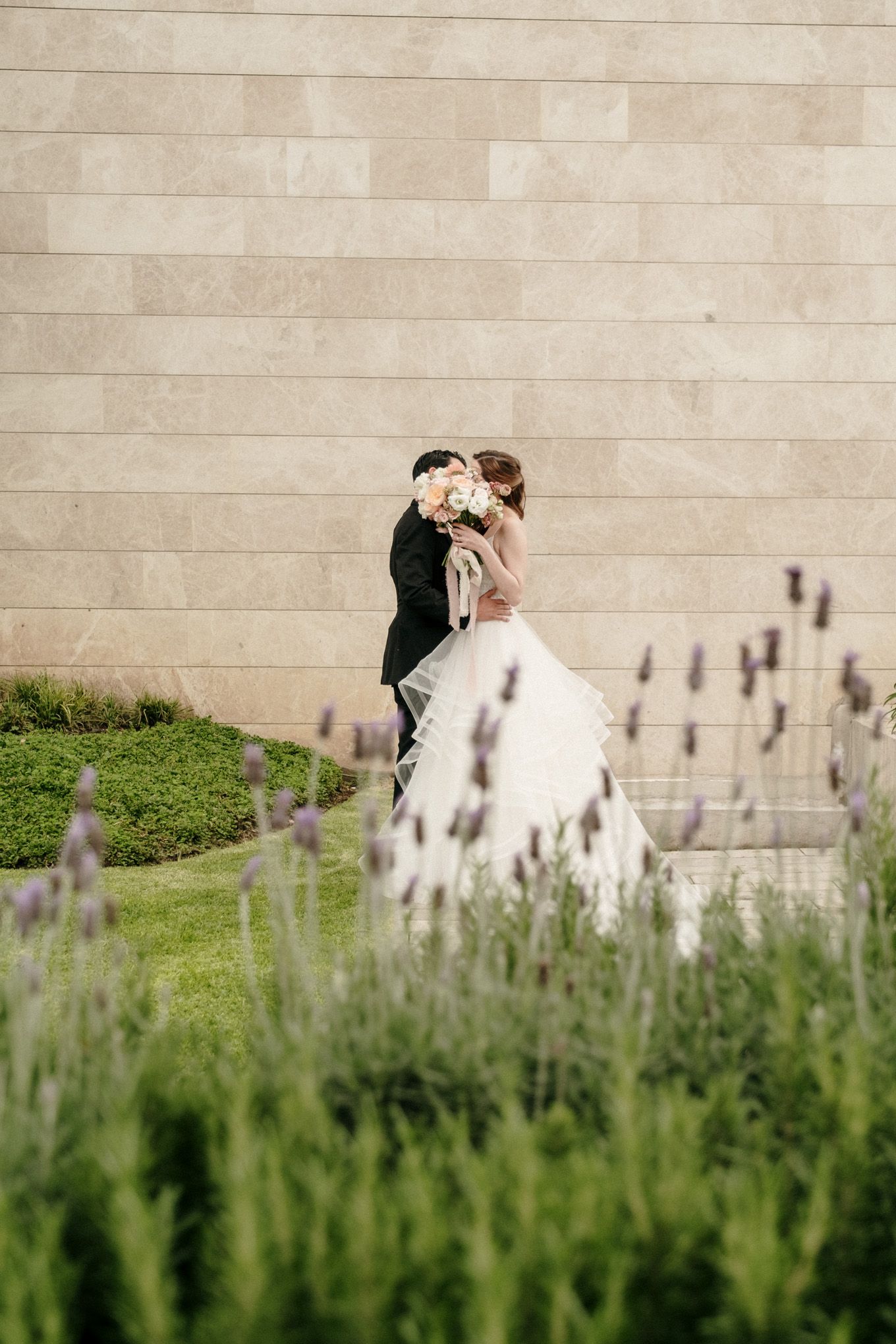 A couple embraces outdoors with a stone wall backdrop and purple lavender flowers in the foreground.
