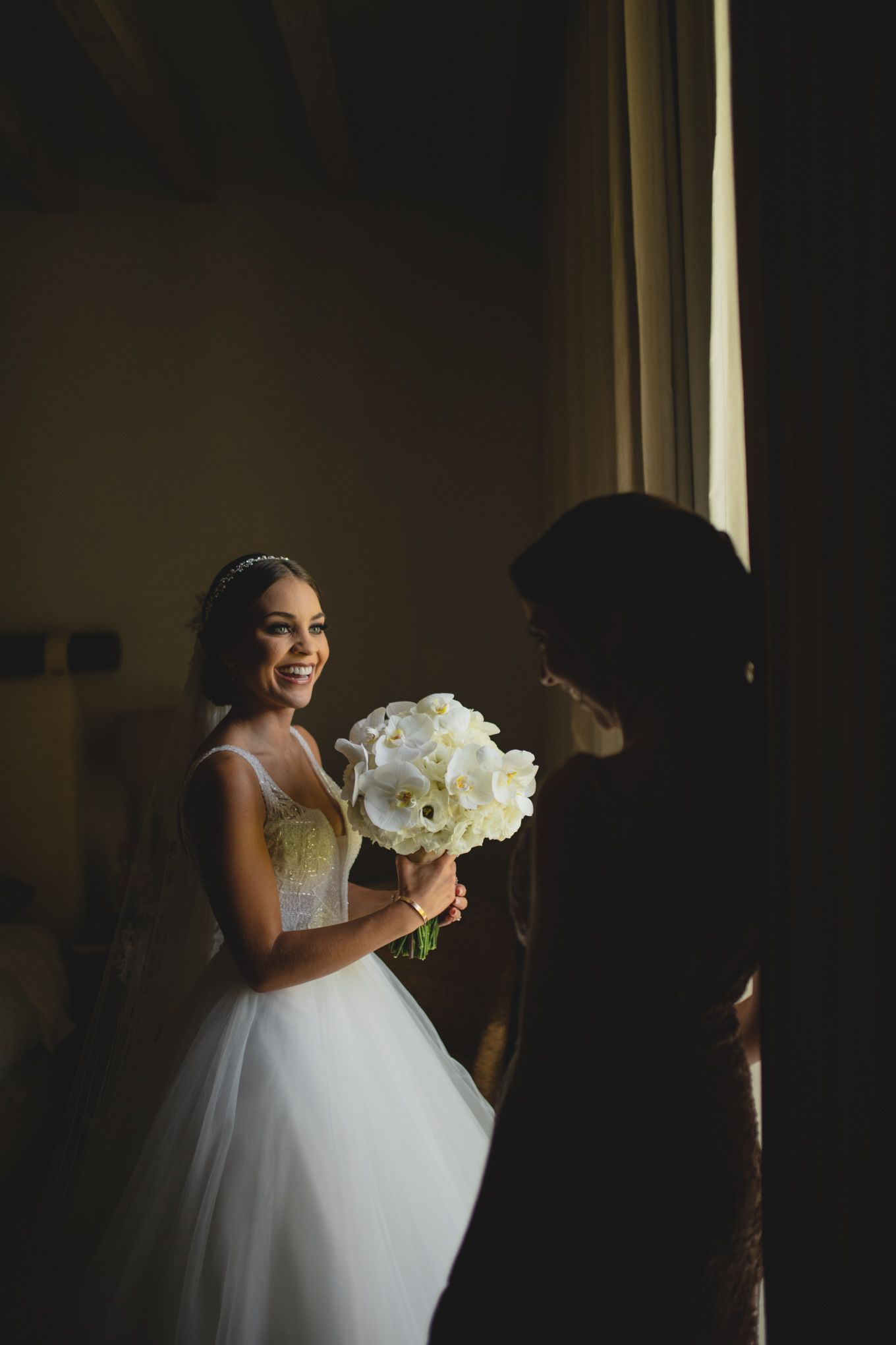 A bride in a white wedding gown stands by a window holding a beautiful bouquet of white calla lilies.