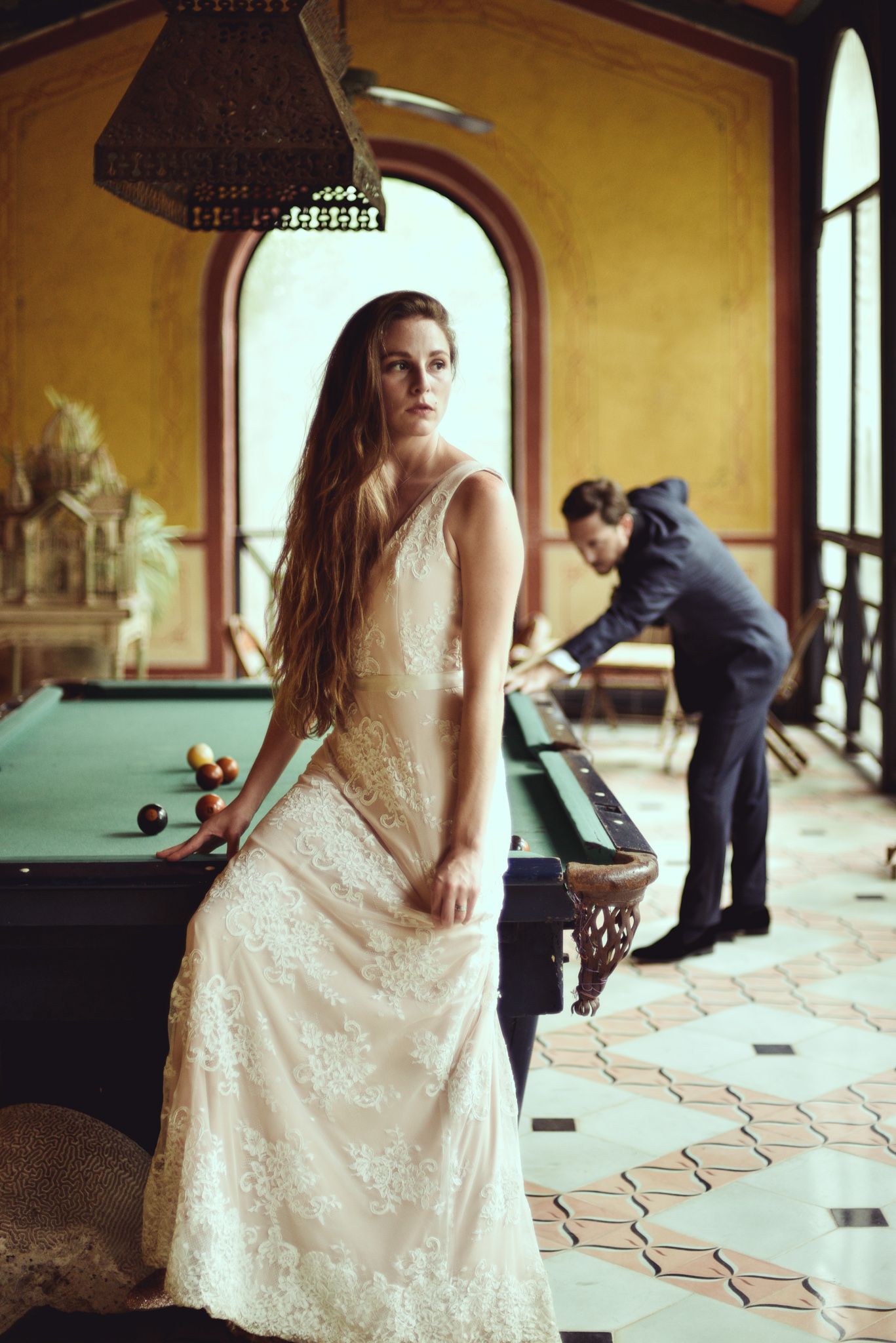 Elegant figure in a lace white dress stands by pool table in vintage room while player leans over table in background.
