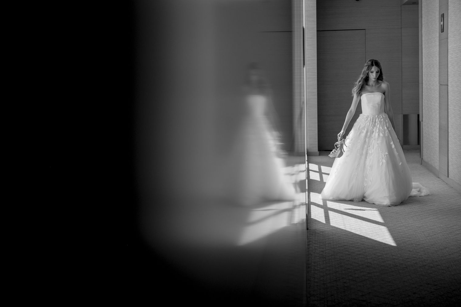 A black and white artistic photo of a bride in a wedding dress standing in dramatic lighting.