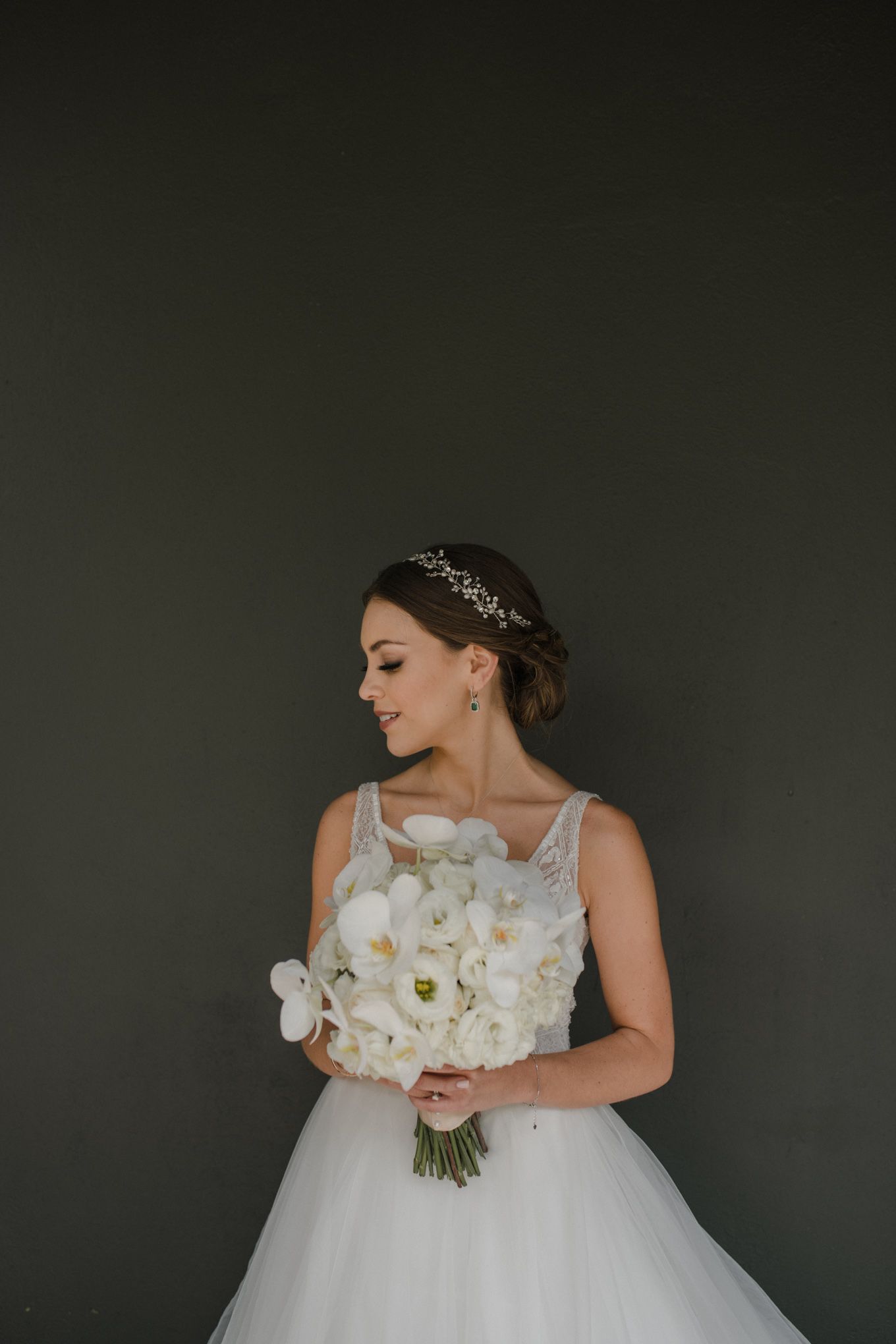 Elegant bride in white gown holds orchid bouquet against dark gray wall.