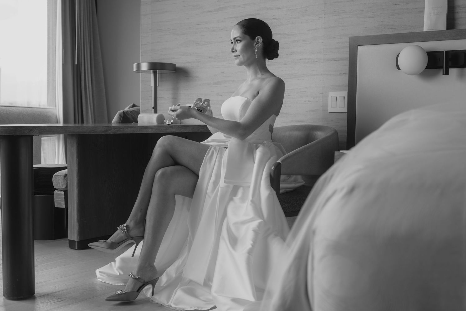 Black and white photo of bride in white dress seated at desk in hotel room.