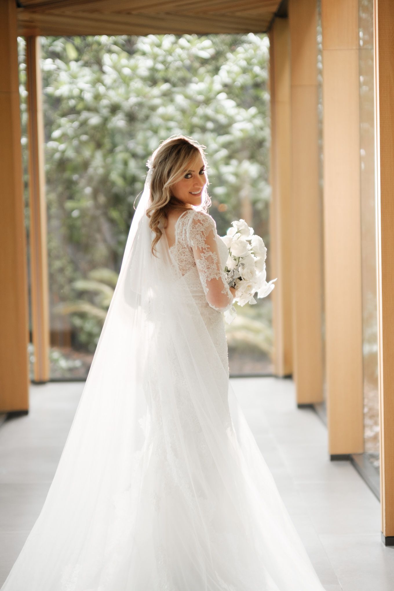 Bride in lace wedding gown with long veil holds white bouquet on covered porch.