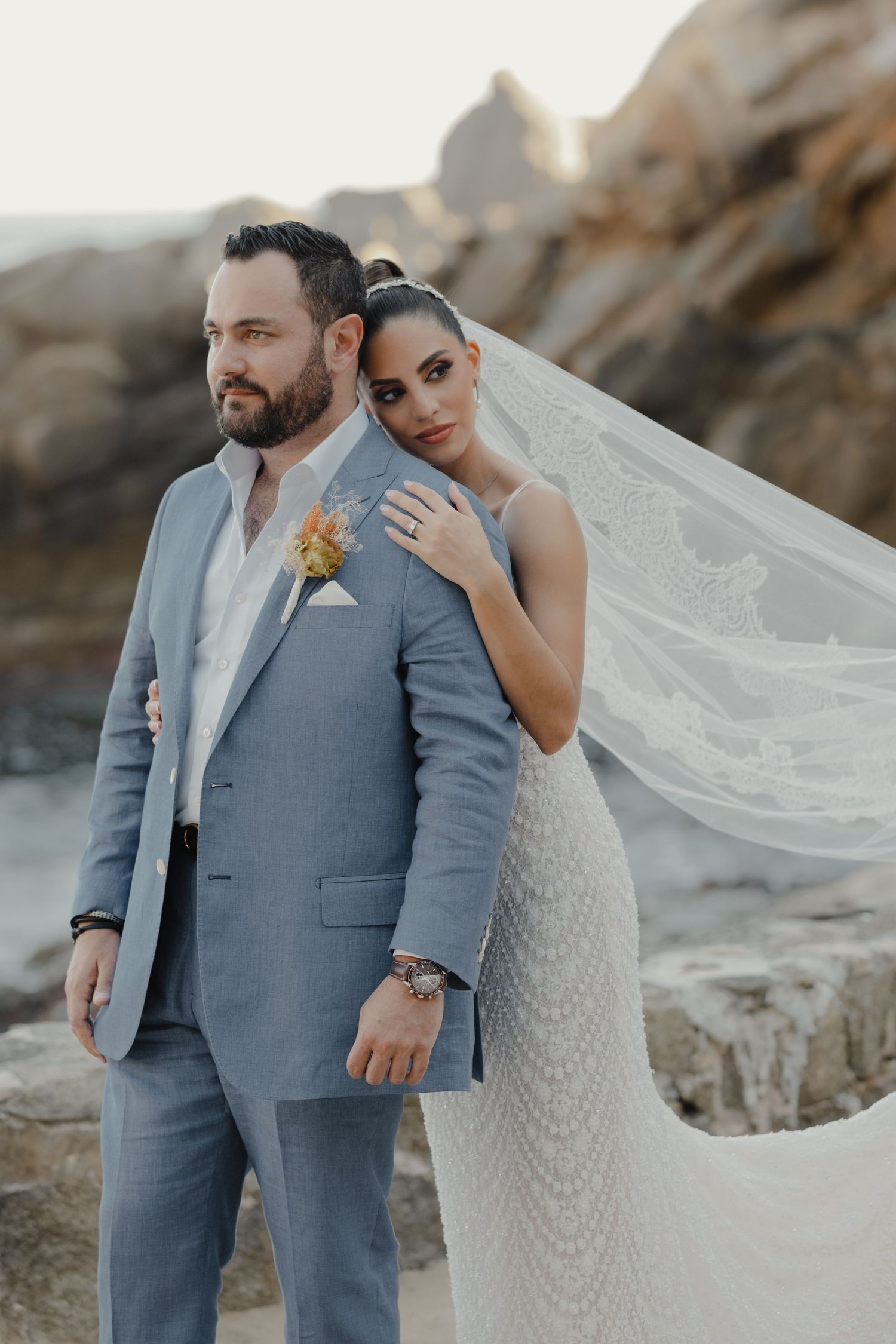 A candid wedding photo with a flowing veil and gray suit against rocky coastal scenery.