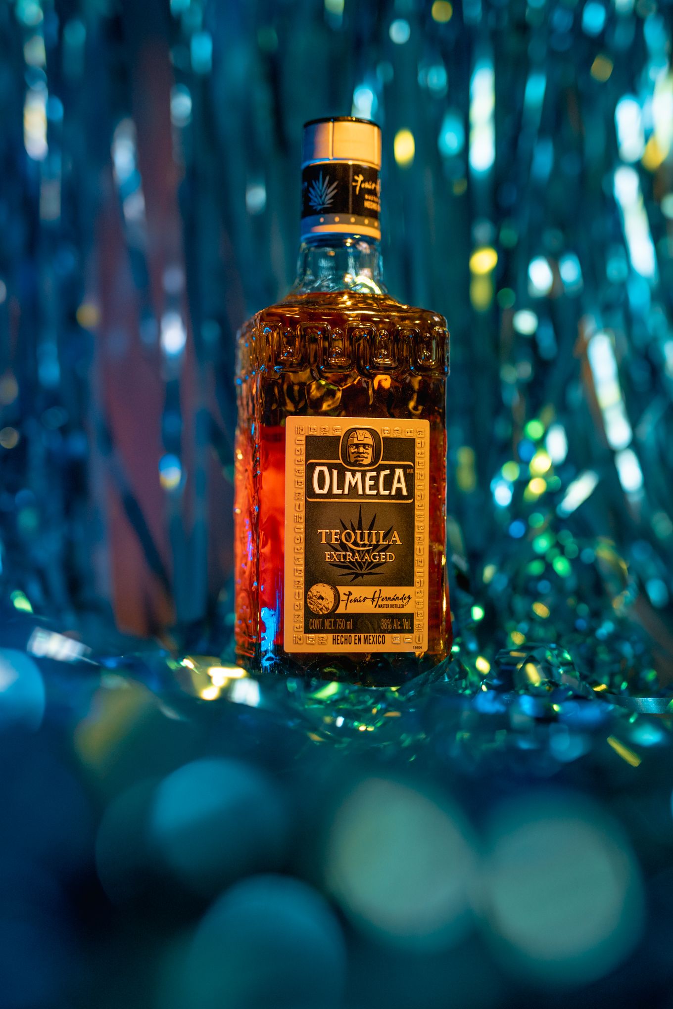 Golden Olmeca tequila bottle showcased against shimmering blue metallic background.
