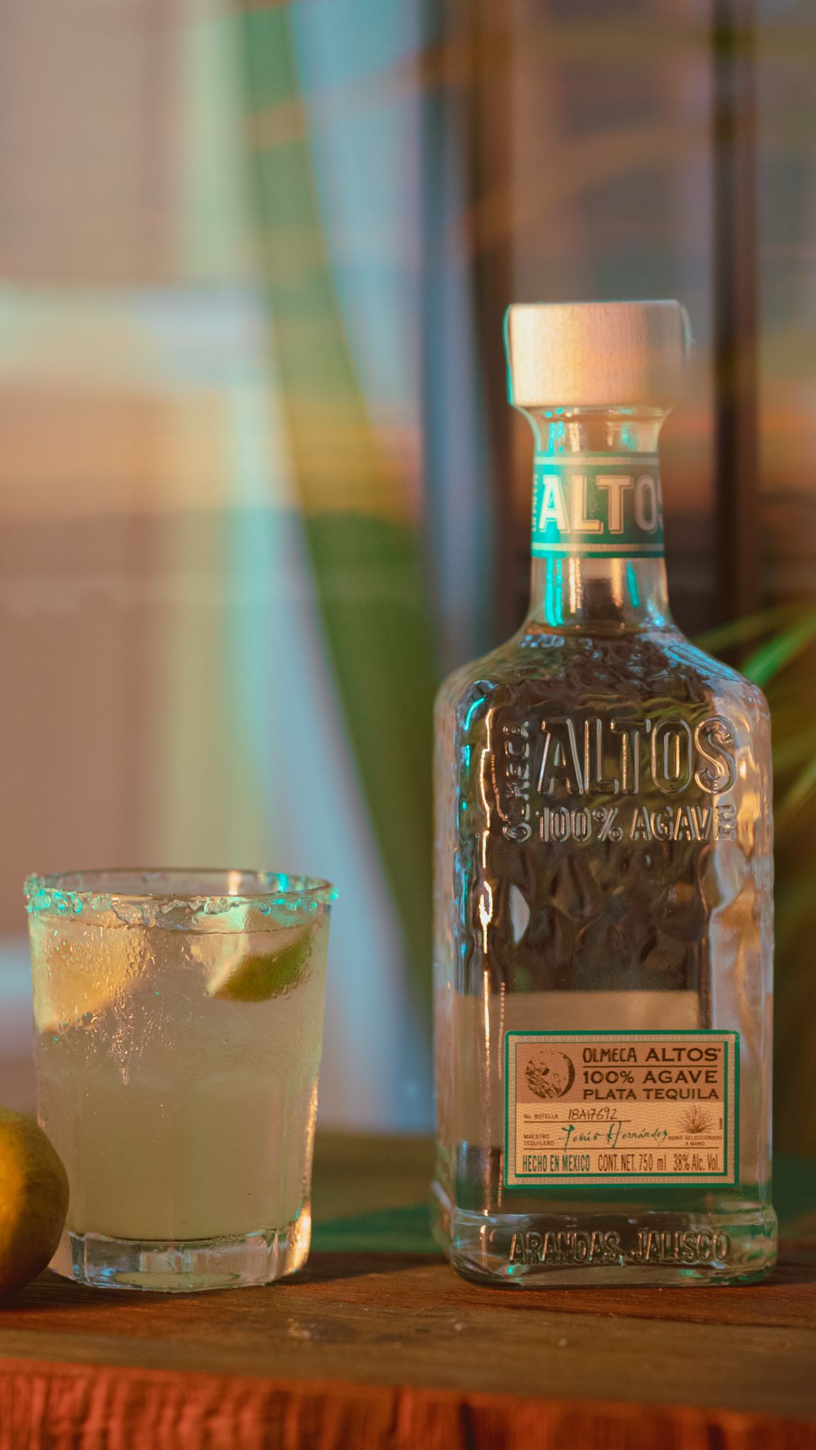 Bottle of Altos tequila next to garnished cocktail on wooden surface.