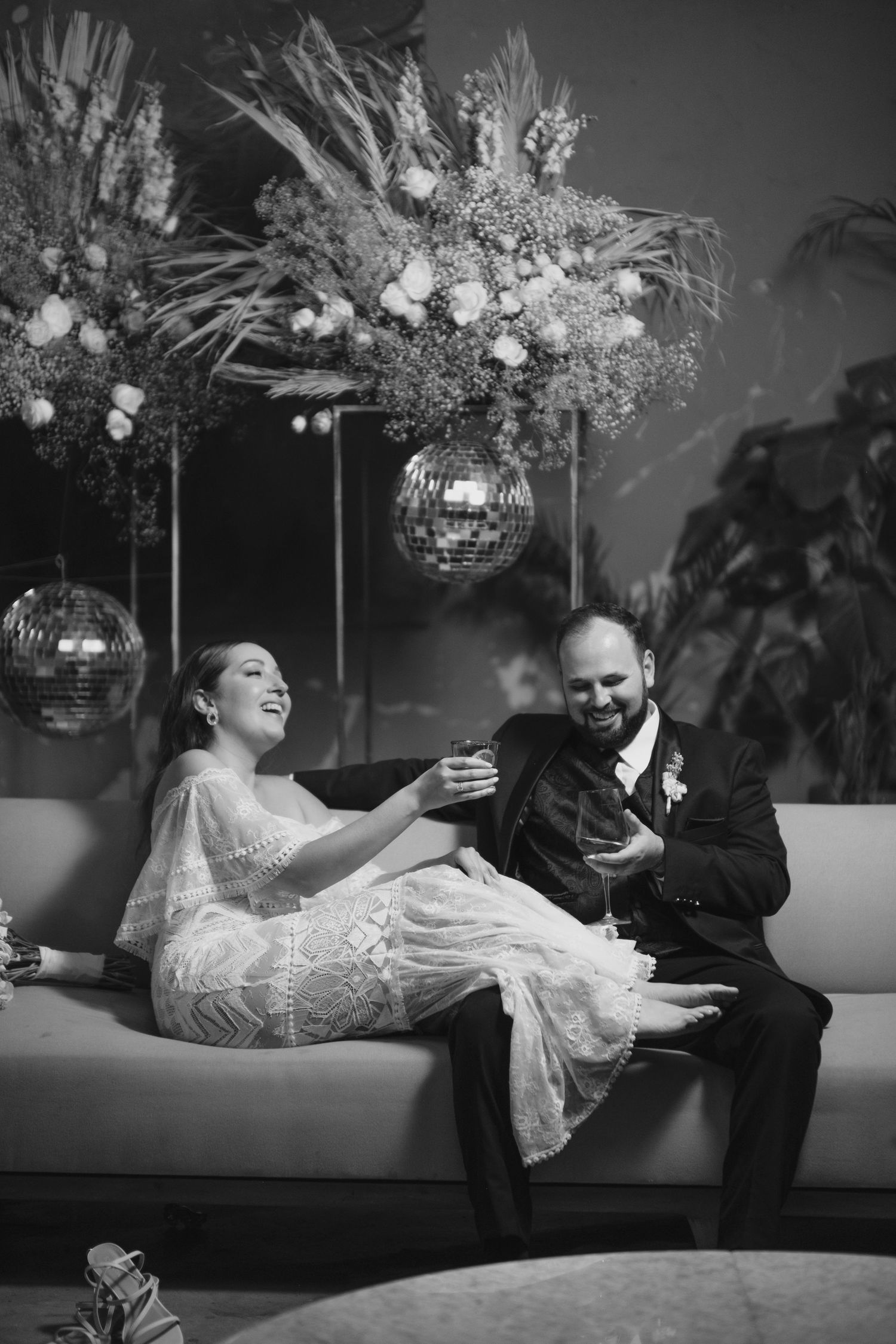 Candid reception moment with couple laughing on couch beneath disco ball and floral arrangements.