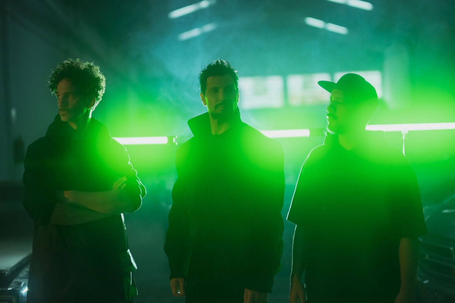 Three silhouetted figures stand against a dramatic green backlit backdrop with fluorescent lights glowing behind them.