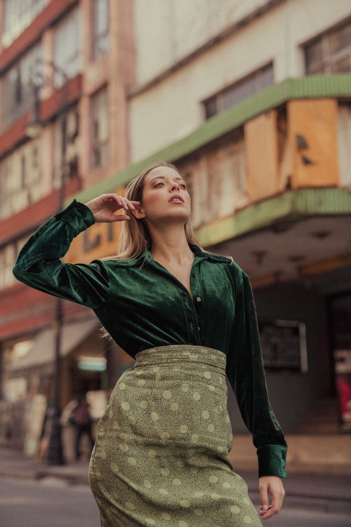 Urban fashion shot featuring green velvet top and patterned skirt against city building.
