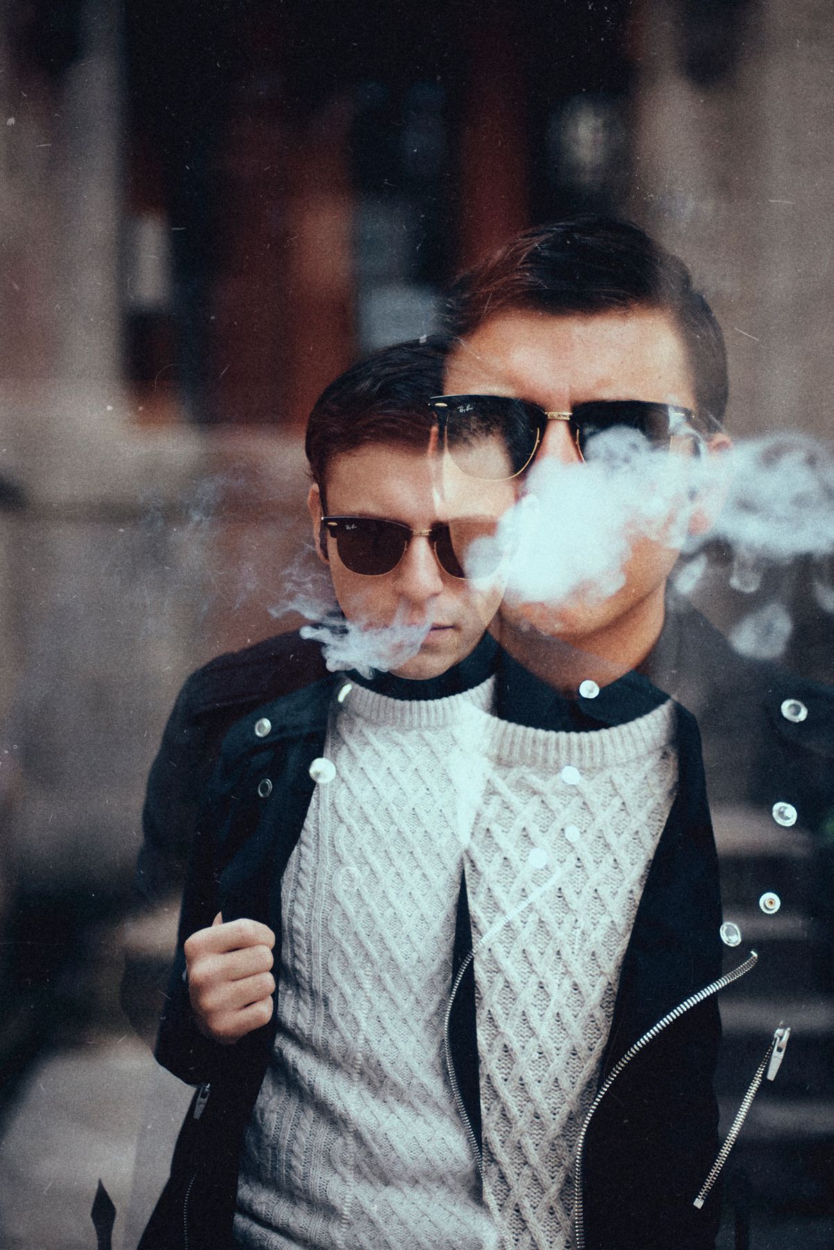 Two people in leather jackets and sunglasses exhaling smoke against urban backdrop.
