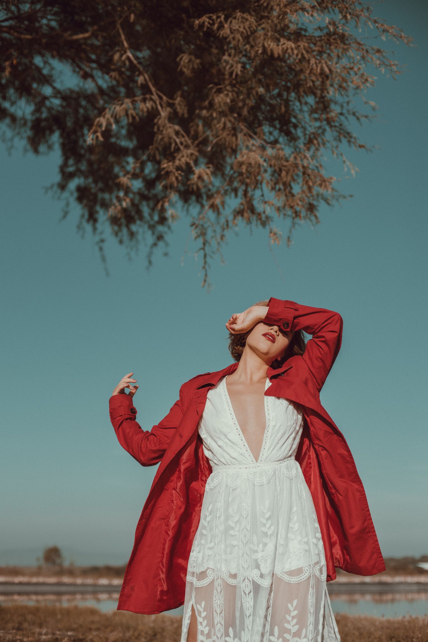 Figure in white dress and red coat poses under tree branch against turquoise sky.