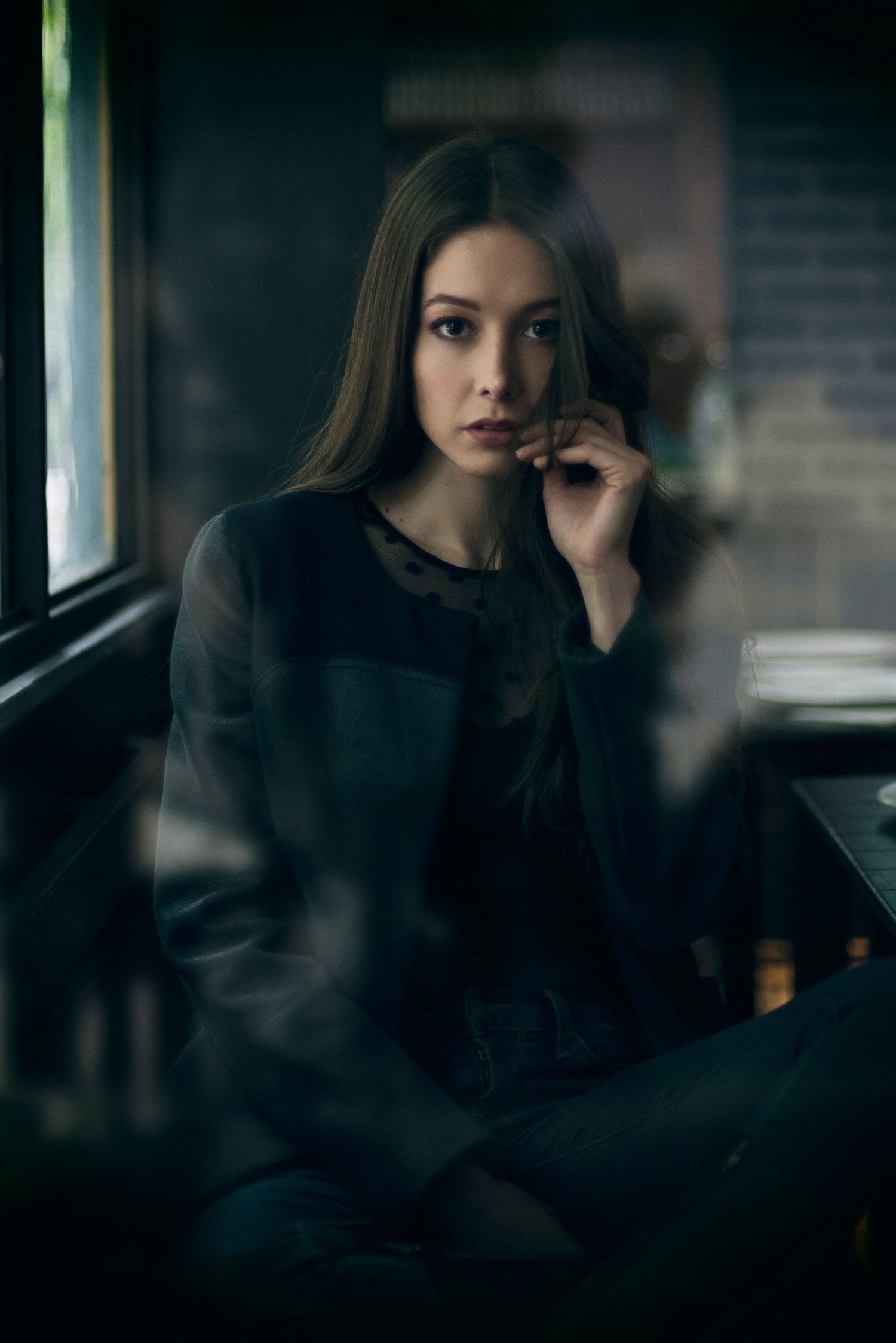 Moody indoor portrait in dark clothing near window with soft natural lighting.