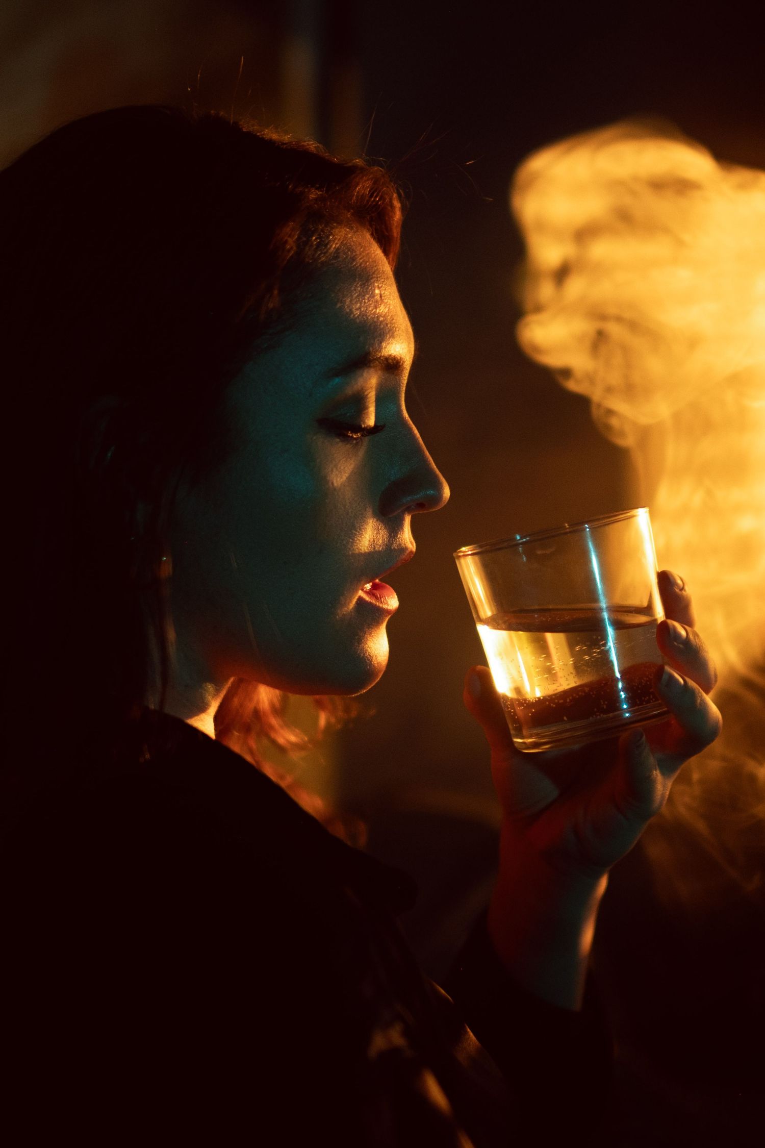 A side profile silhouetted against orange light holds a glass of whiskey while smoke curls in the background.