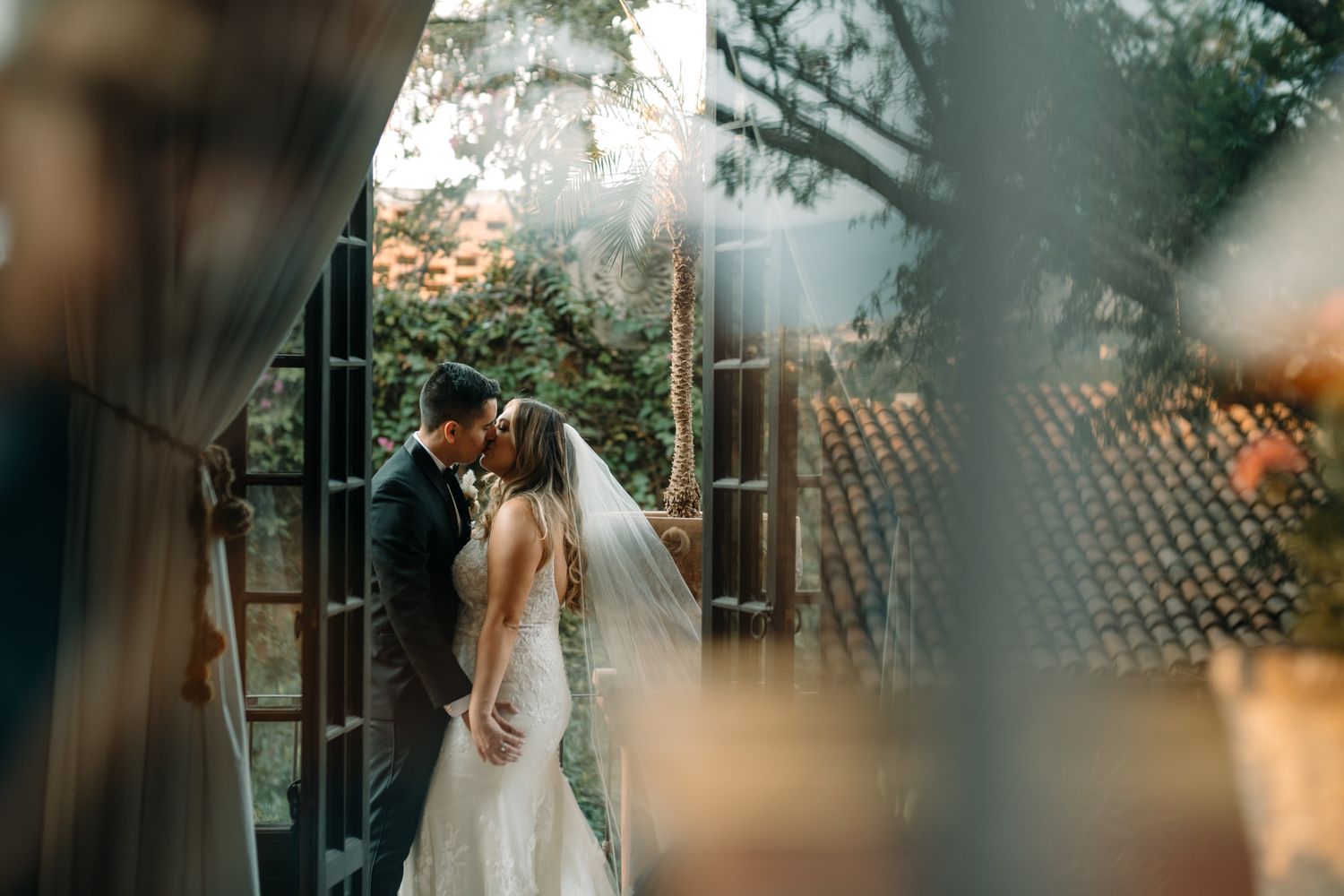 Romantic wedding moment captured through blurred foreground elements in a garden setting.