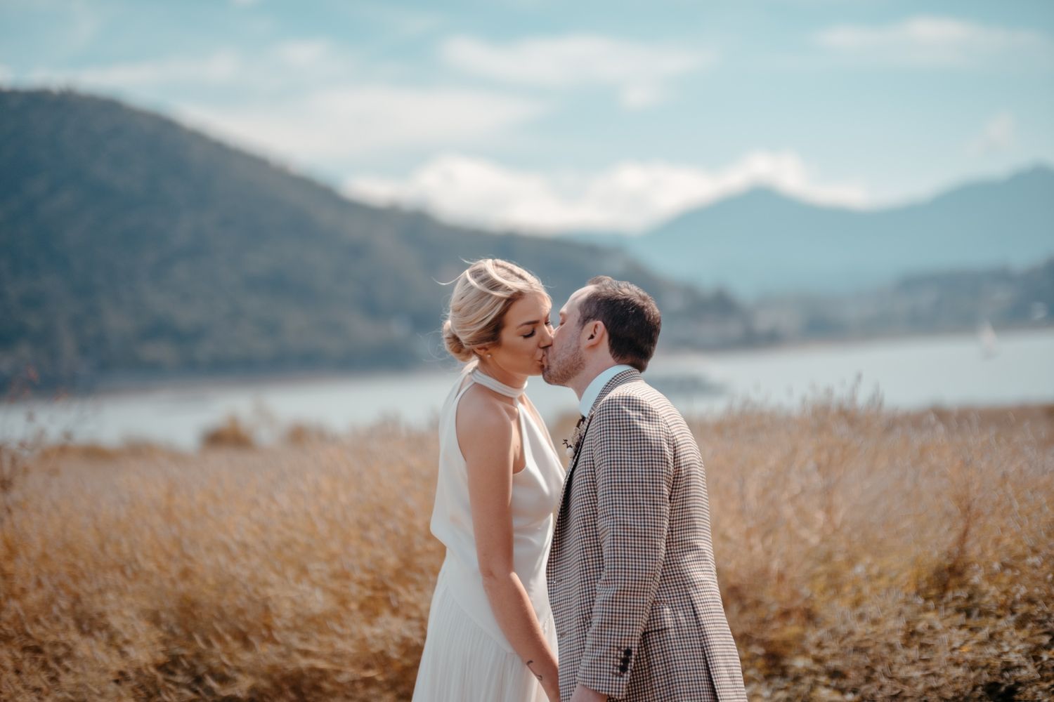 Wedding couple embracing in golden field with mountains and lake in background.