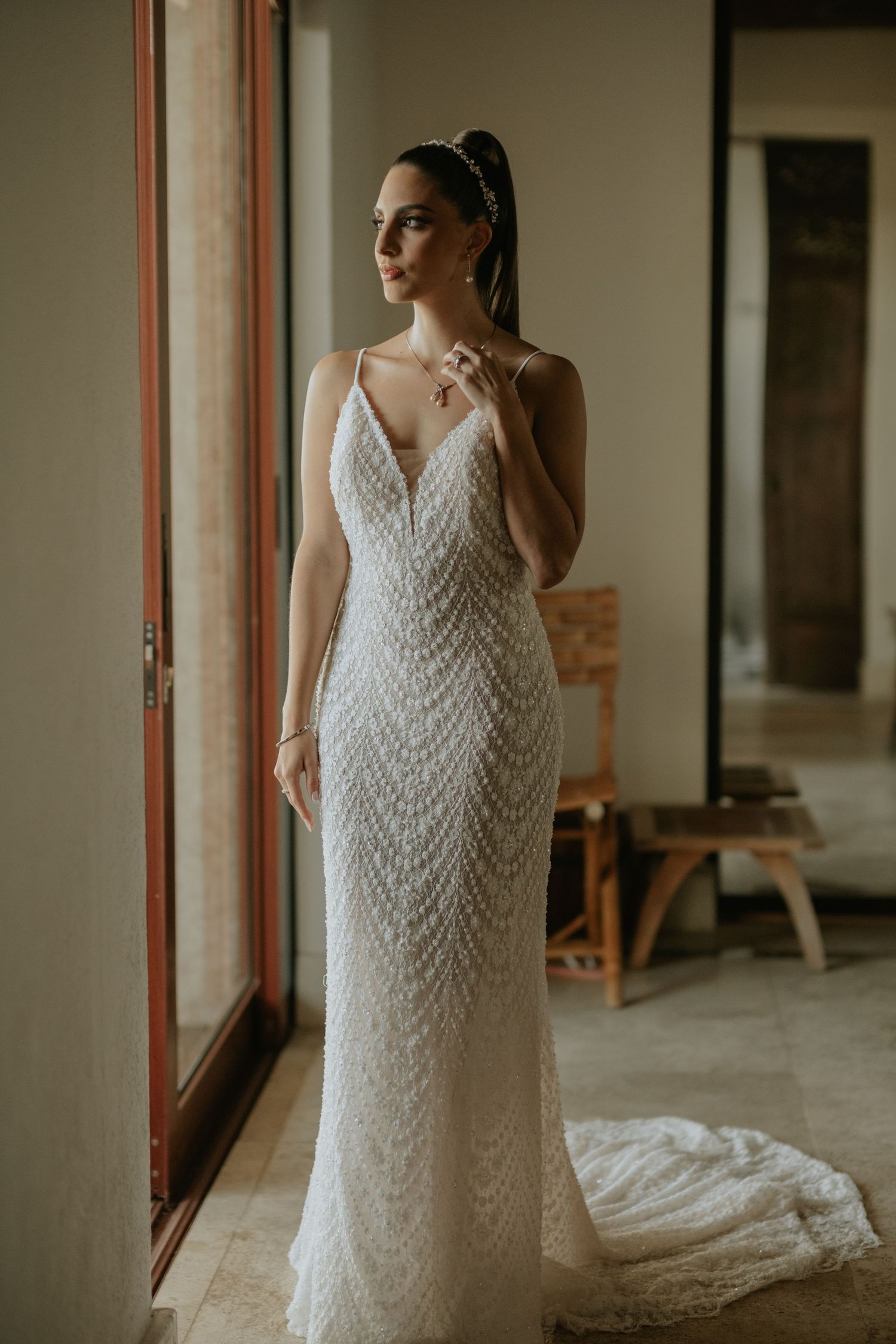 Bride in fitted chevron lace wedding dress stands by window in natural light.
