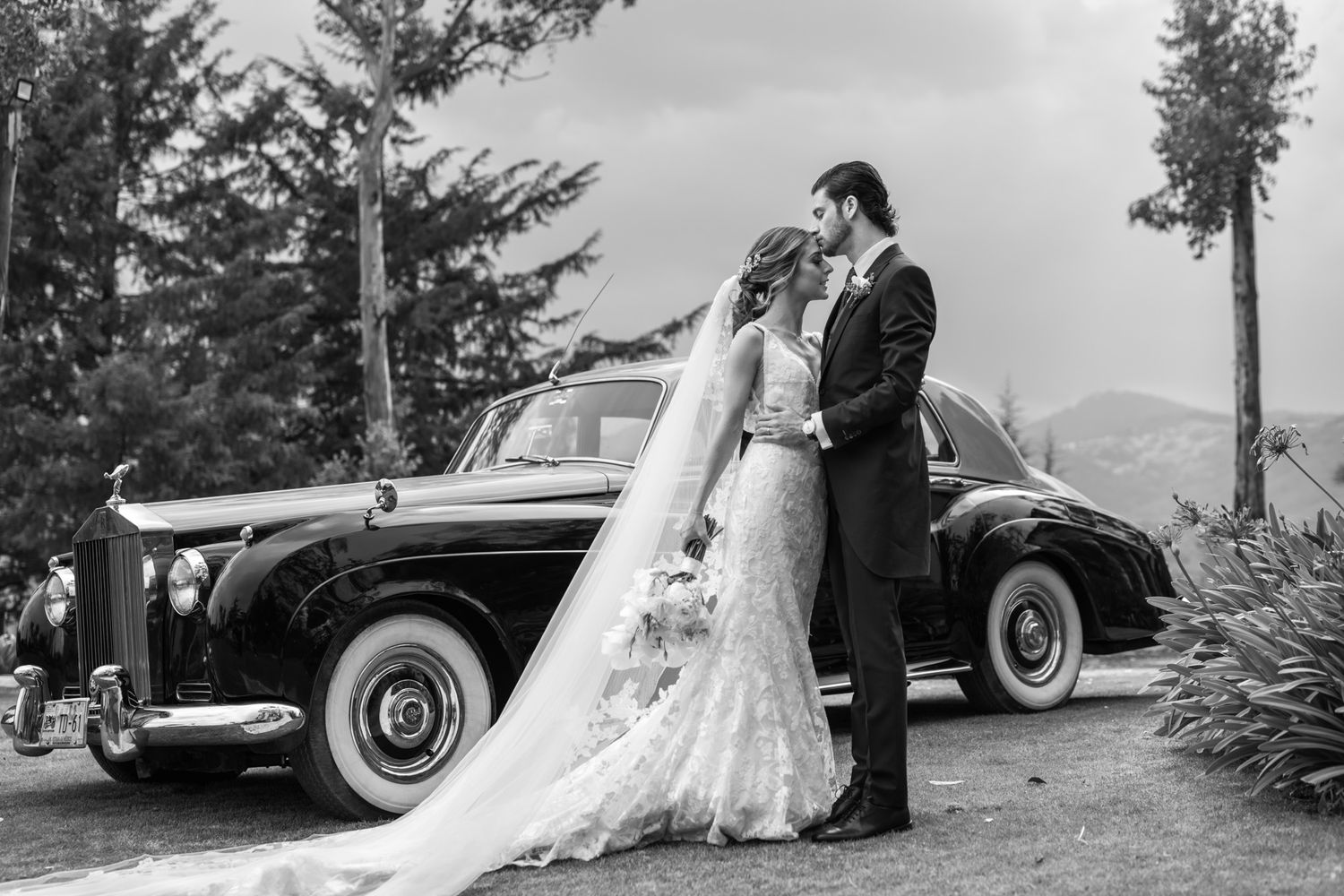 Black and white wedding portrait with couple posing beside vintage luxury car.