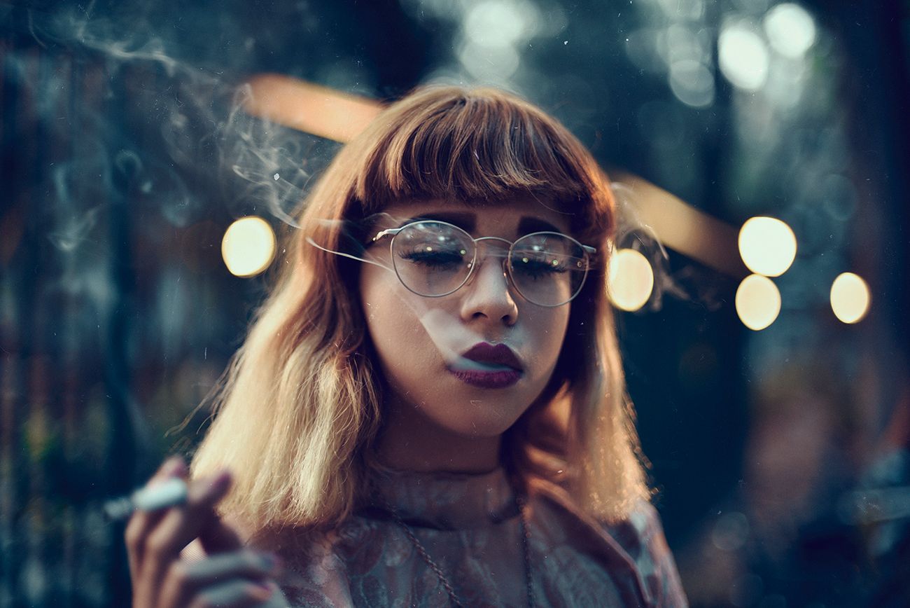 Artistic portrait of person with bangs smoking while wearing glasses in moody lighting.