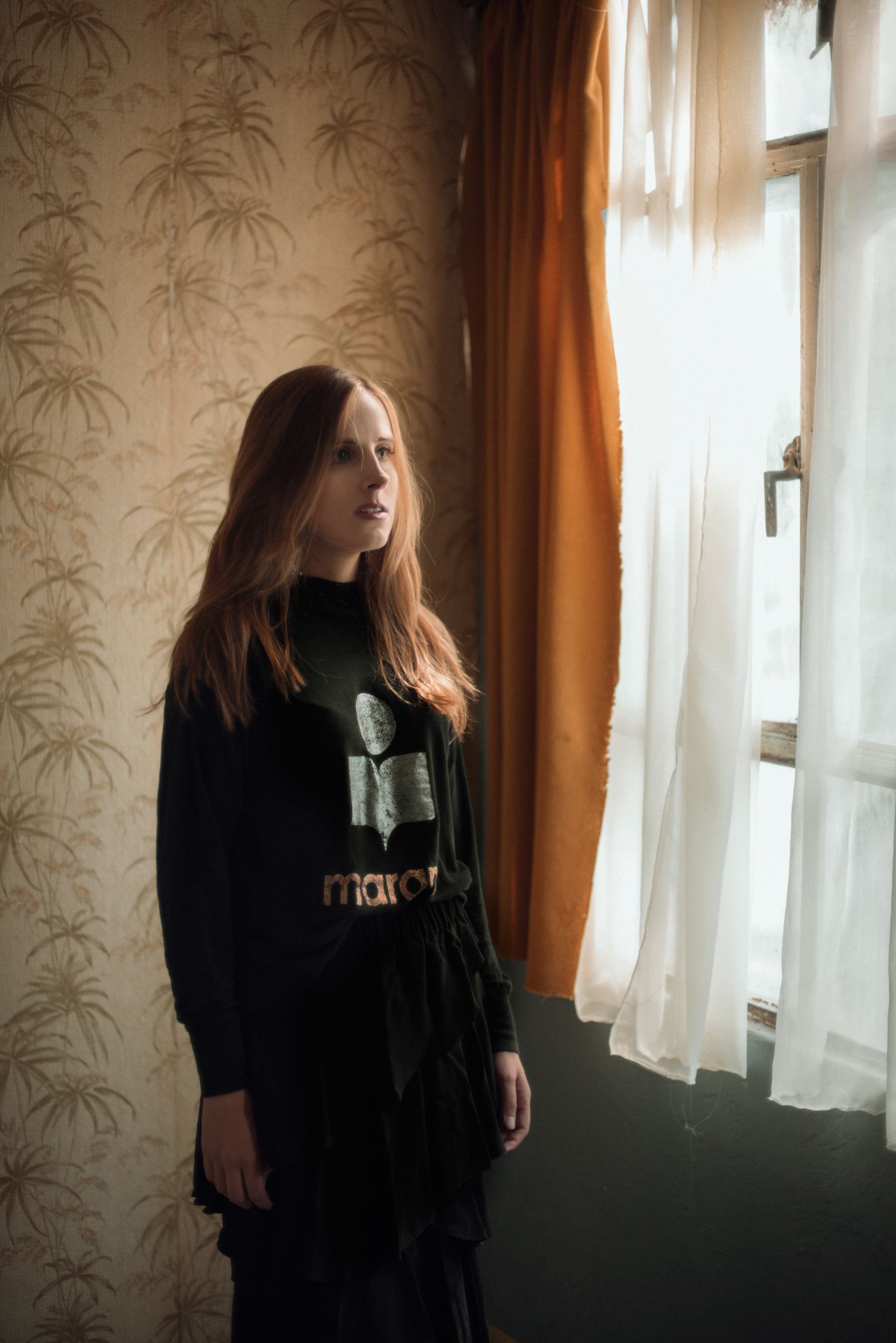 Moody indoor portrait by window with palm print wallpaper and orange curtains.