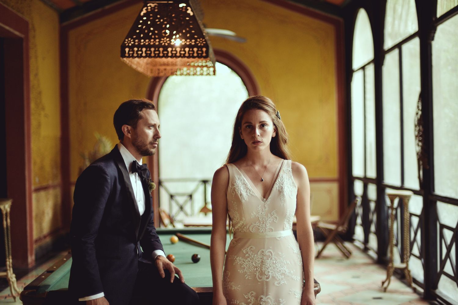 Elegant couple in formal attire standing near large arched windows in a vintage interior setting with ornate lighting.