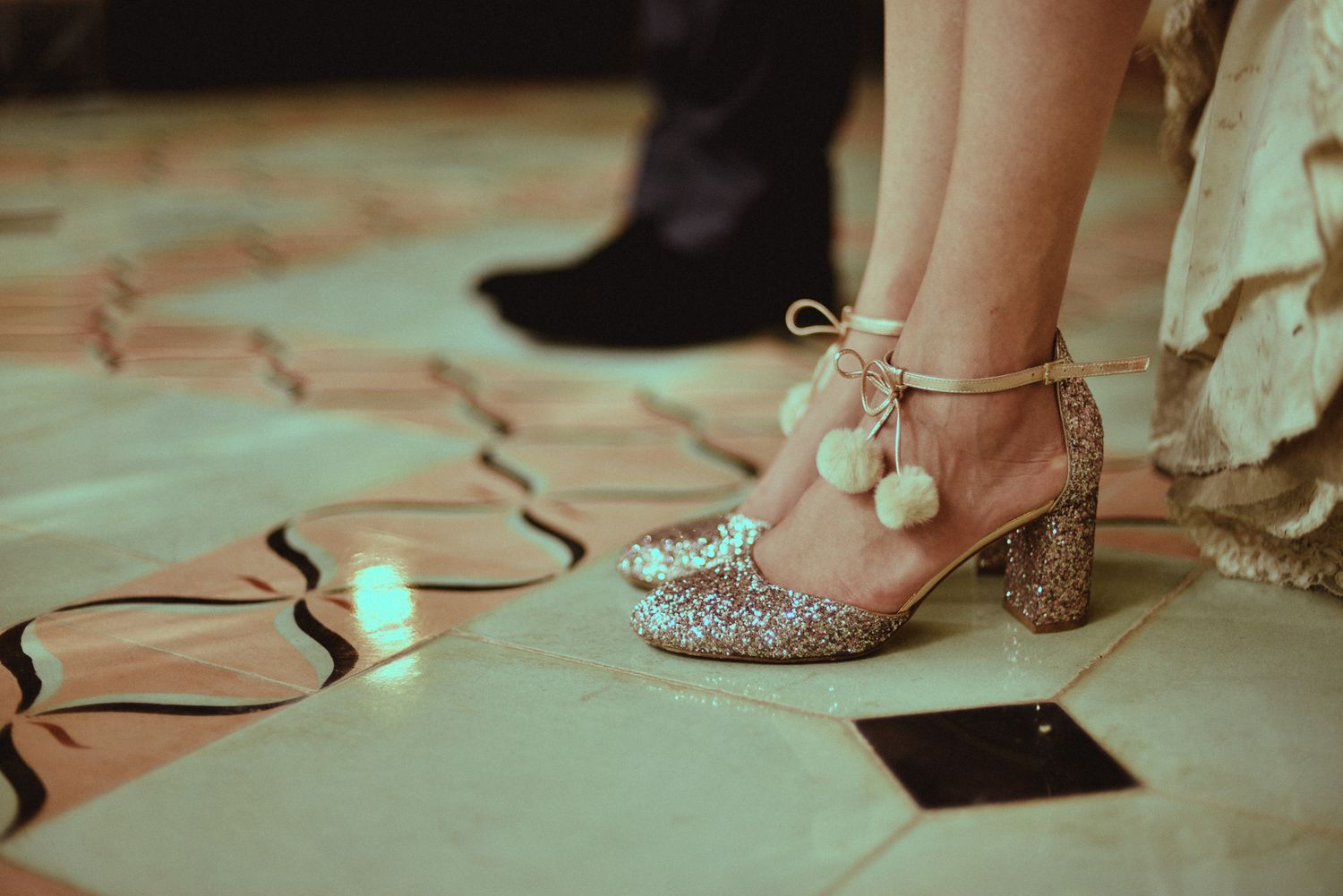 Sparkly silver heels with pom pom ties stand out on a geometric patterned dance floor with scattered confetti.