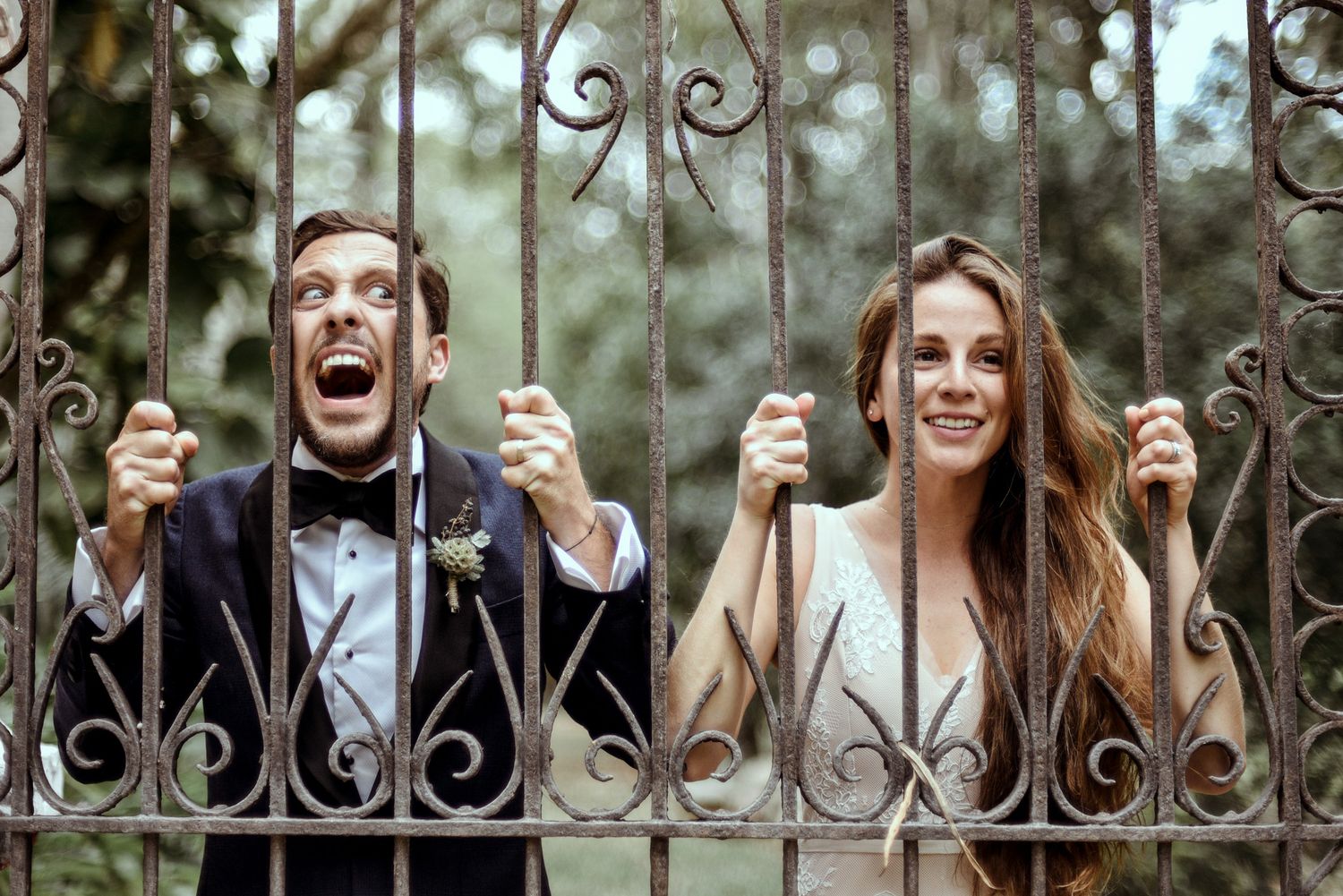 Happy newlyweds peek through ornate iron gates while playfully holding the bars and sharing a joyful moment together.