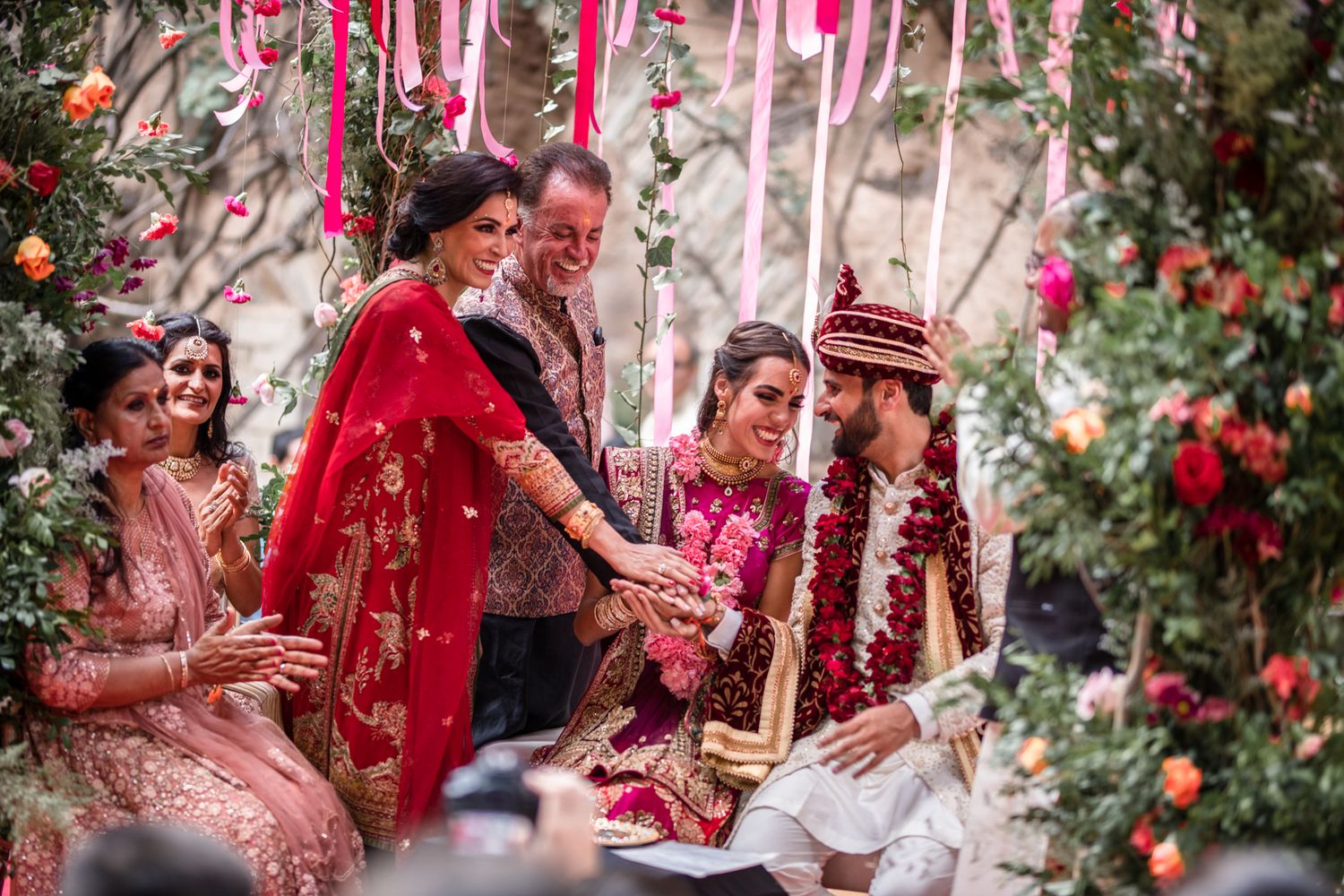 Traditional Indian wedding ceremony with bright red and pink decorations, floral arrangements, and flowing ribbons in an outdoor setting.