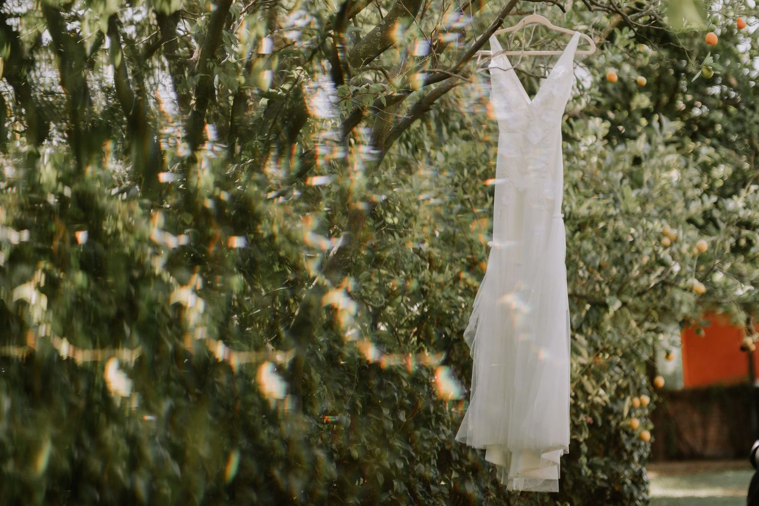 A white wedding dress hangs elegantly from a hanger outdoors with twinkling lights and greenery in the background.