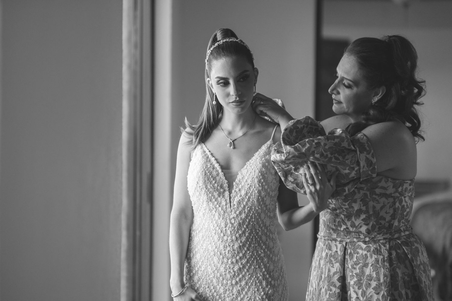 A black and white wedding preparation moment showing final adjustments being made to an ornate lace gown.