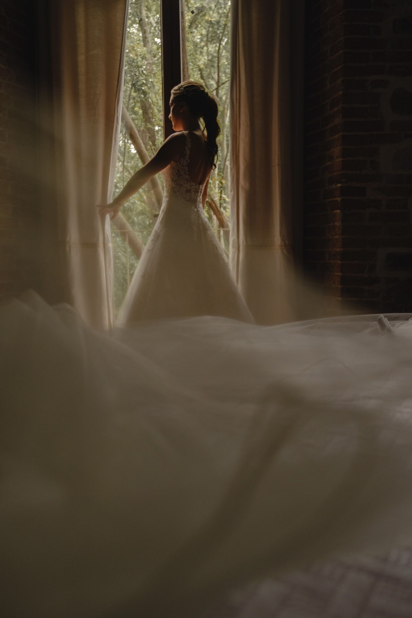 Silhouette of a figure in a flowing white wedding dress standing by a sunlit window with sheer curtains.
