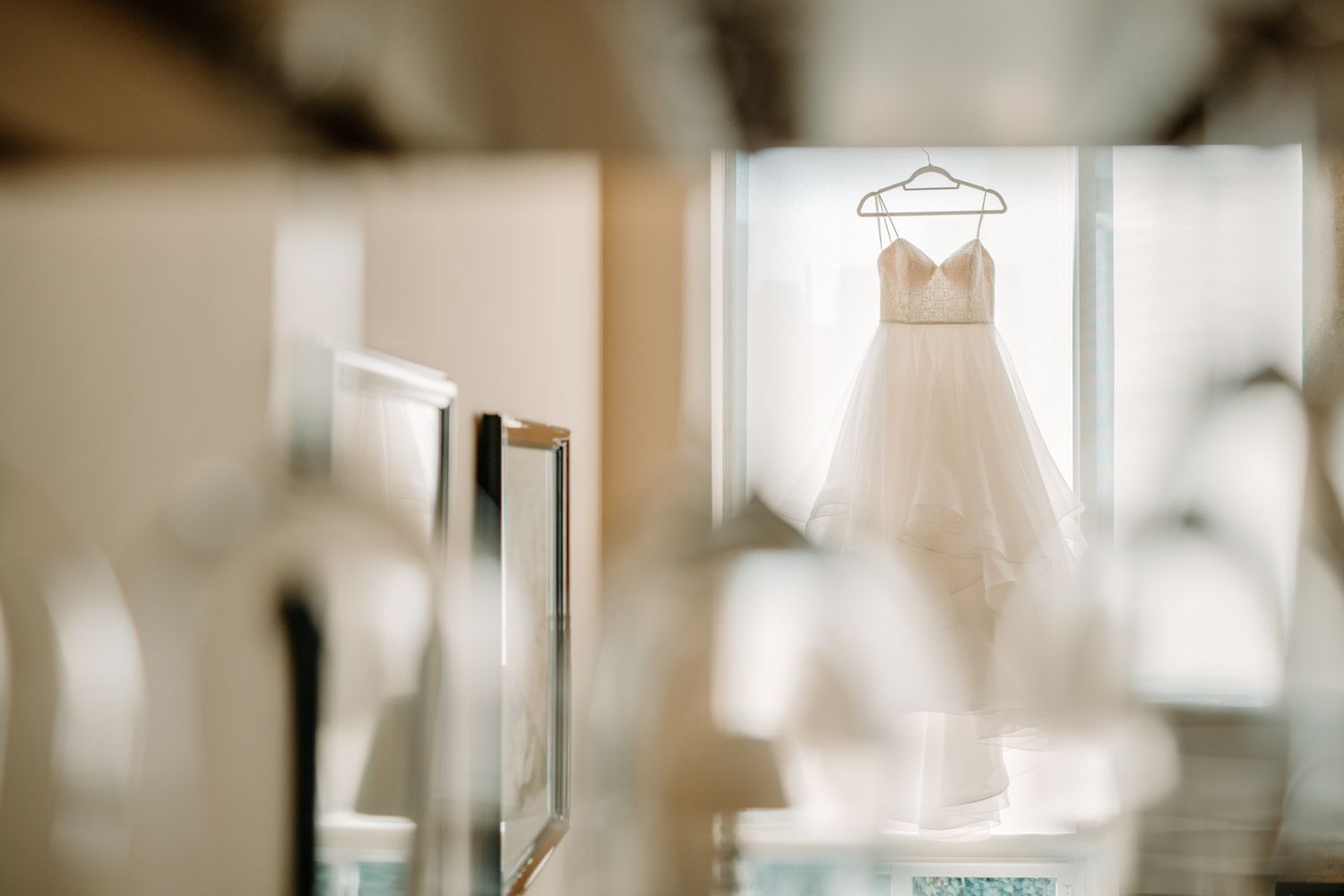 A white wedding dress hangs illuminated in front of a window creating a dreamy silhouette in soft natural light.